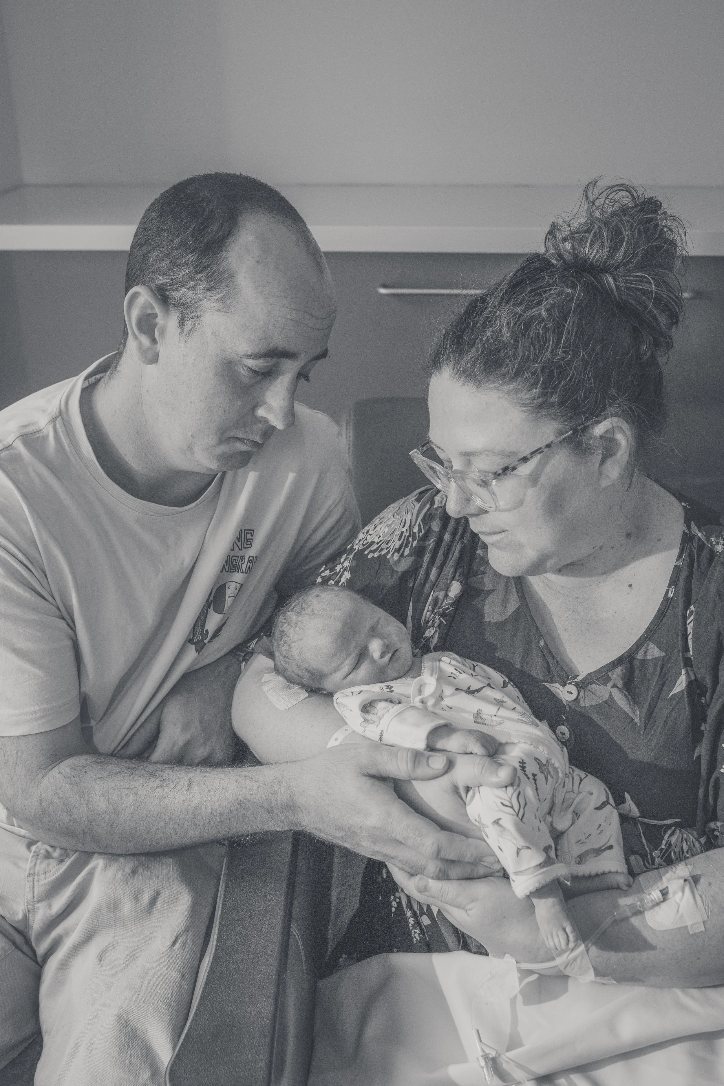 A man and woman holding a newborn baby at a hospital, with medical equipment visible.