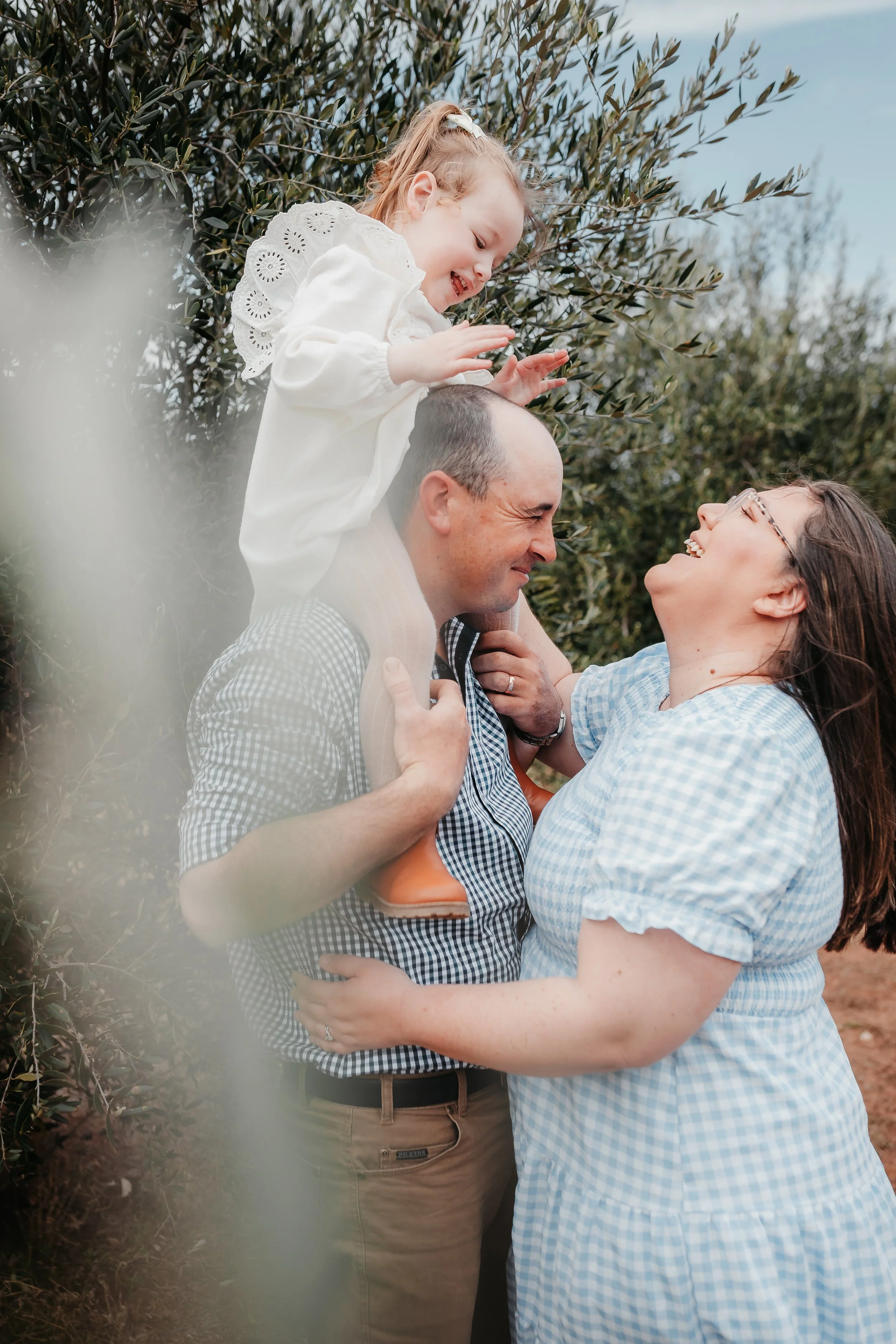 A family of three is outside among trees, with a woman and man holding a smiling young girl on their shoulders. They are enjoying a joyful moment together.