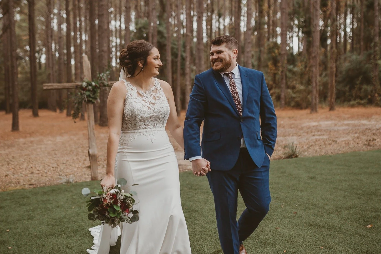 A bride and groom walking hand in hand outdoors in a forested area during their wedding, smiling at each other.