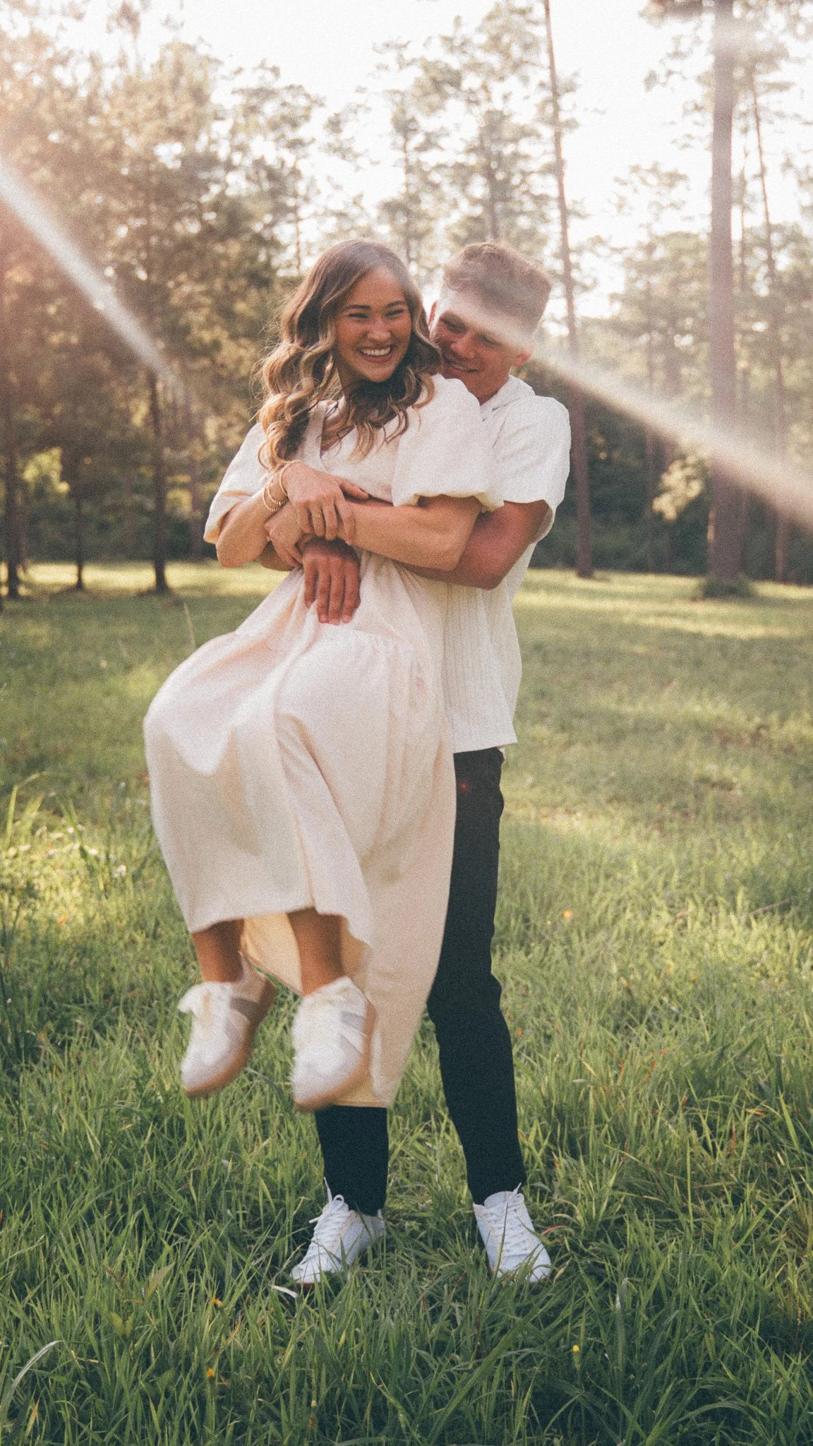 A young couple is smiling and hugging outdoors in a grassy field with trees in the background. The woman is being lifted by the man, and they both appear happy and joyful. The sunlight creates a warm, glowing atmosphere.
