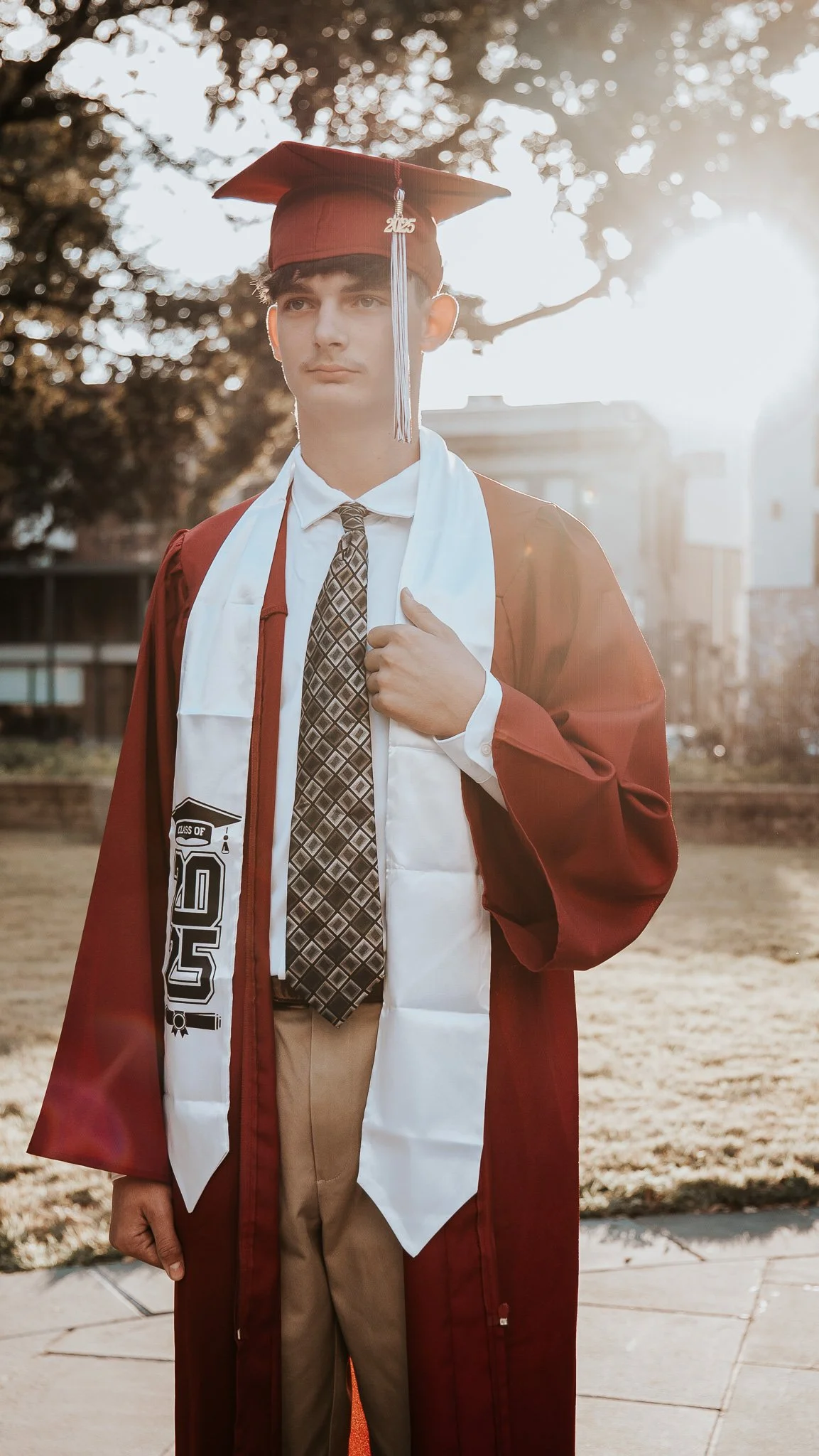 A young man in a maroon graduation cap and gown, with a white stole and a patterned tie, standing outdoors during sunset, holding his gown with one hand.