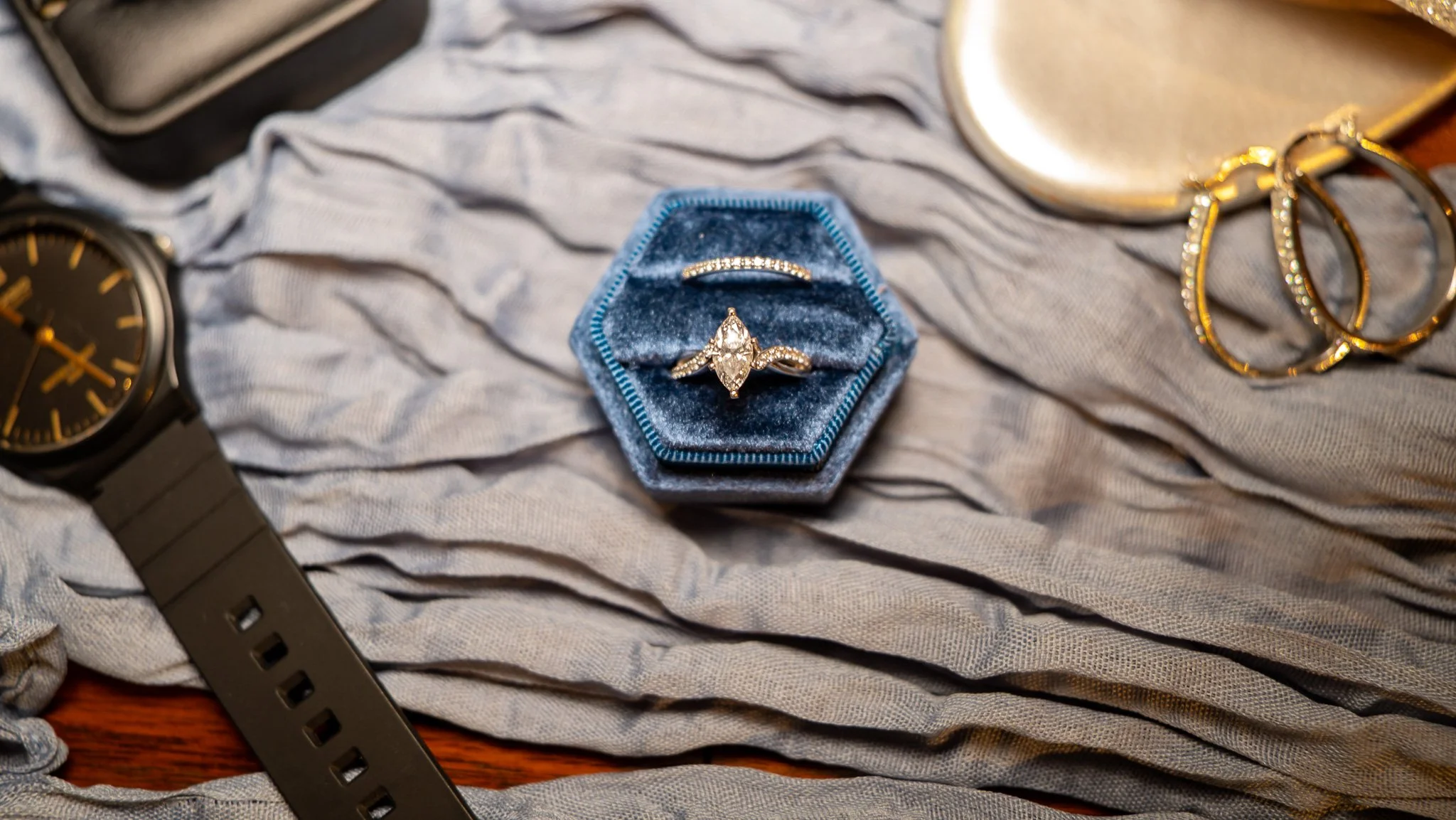 A diamond engagement ring in a blue velvet ring box, alongside gold hoop earrings, a black watch, and a gold clutch purse, all arranged on a gray fabric background.