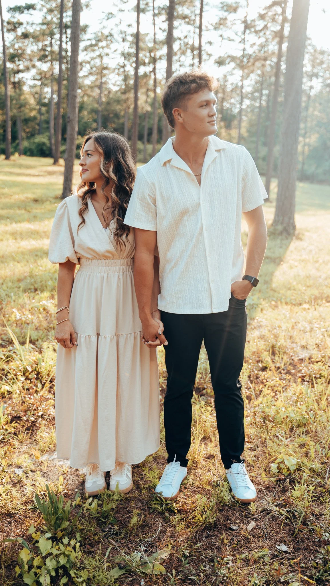 A young couple holding hands outdoors in a forested area during golden hour, with trees and sunlight in the background.