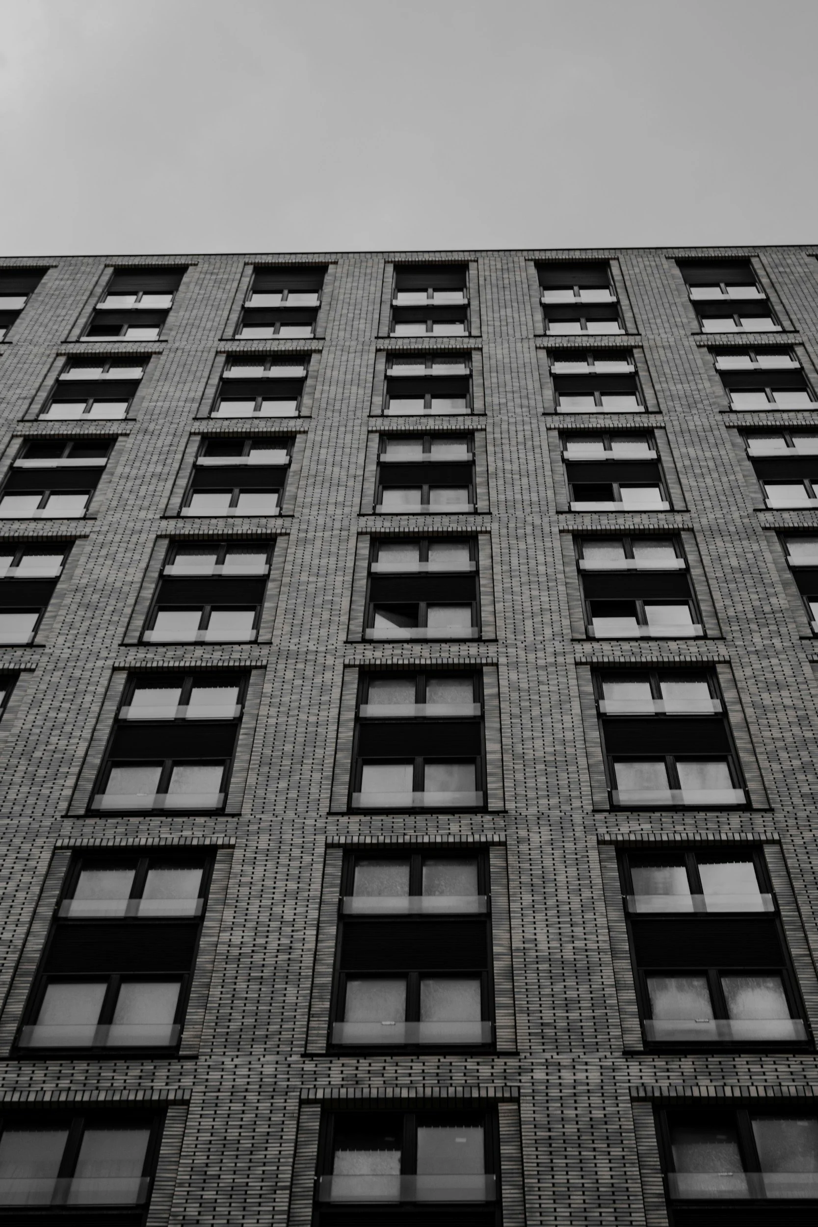 A black-and-white photo of a tall apartment or office building with multiple windows, taken from a low angle with a cloudy sky above.