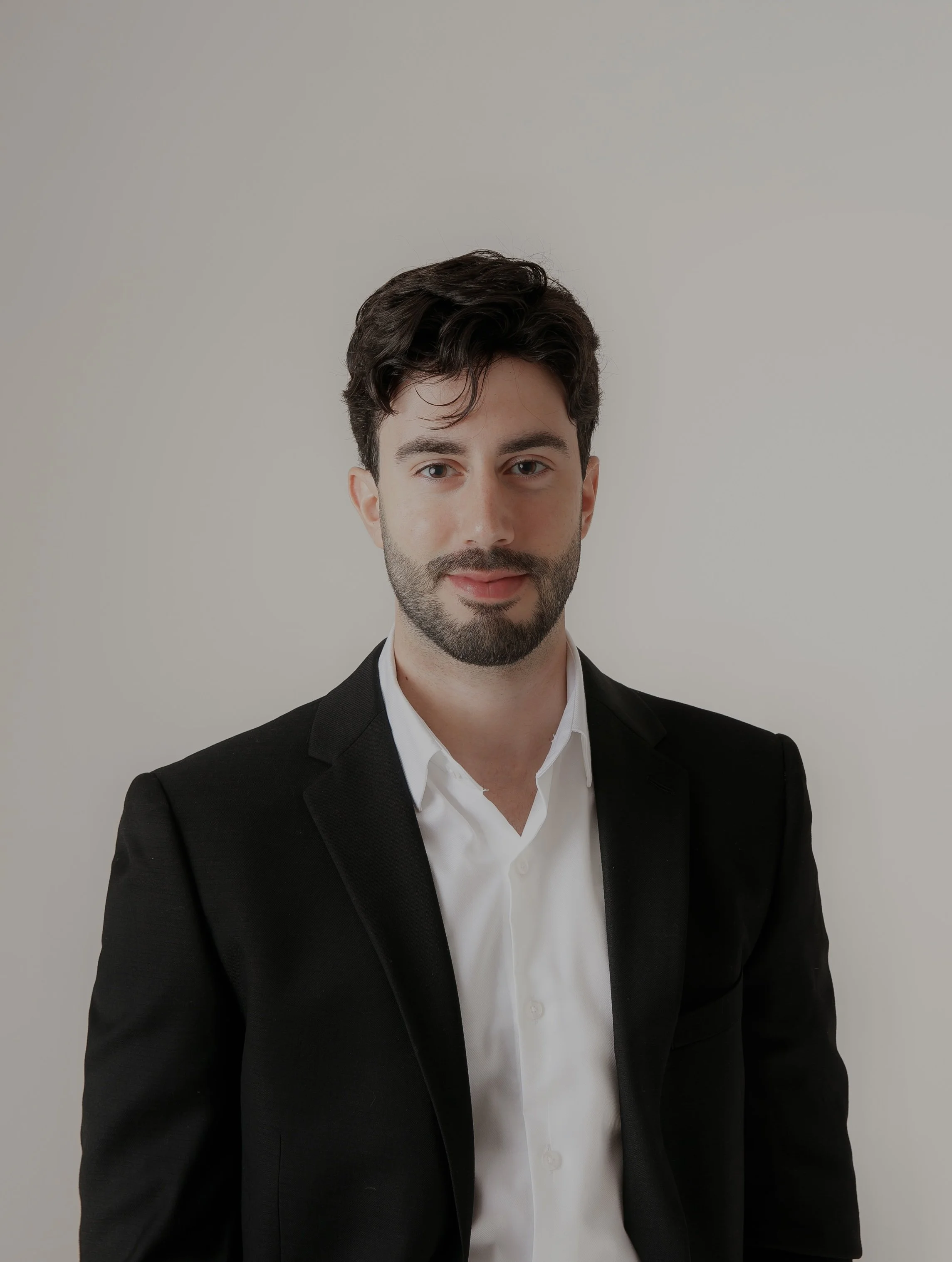 A young man with dark hair and a beard wearing a black suit jacket and a white dress shirt standing against a plain light grey background.