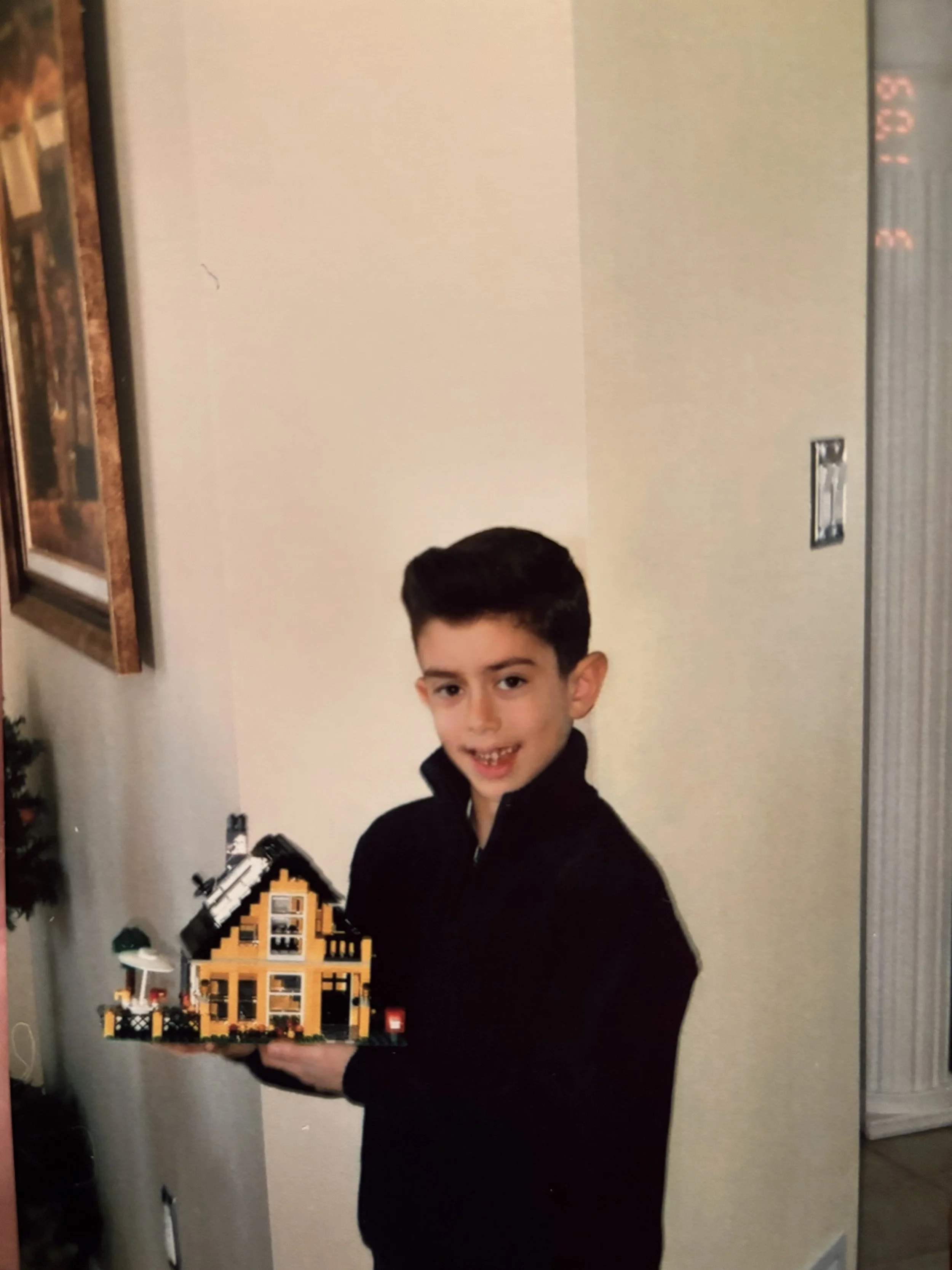 A young boy with dark hair and wearing a black jacket smiling and holding a LEGO house with yellow walls and black roof in front of a plain beige wall.