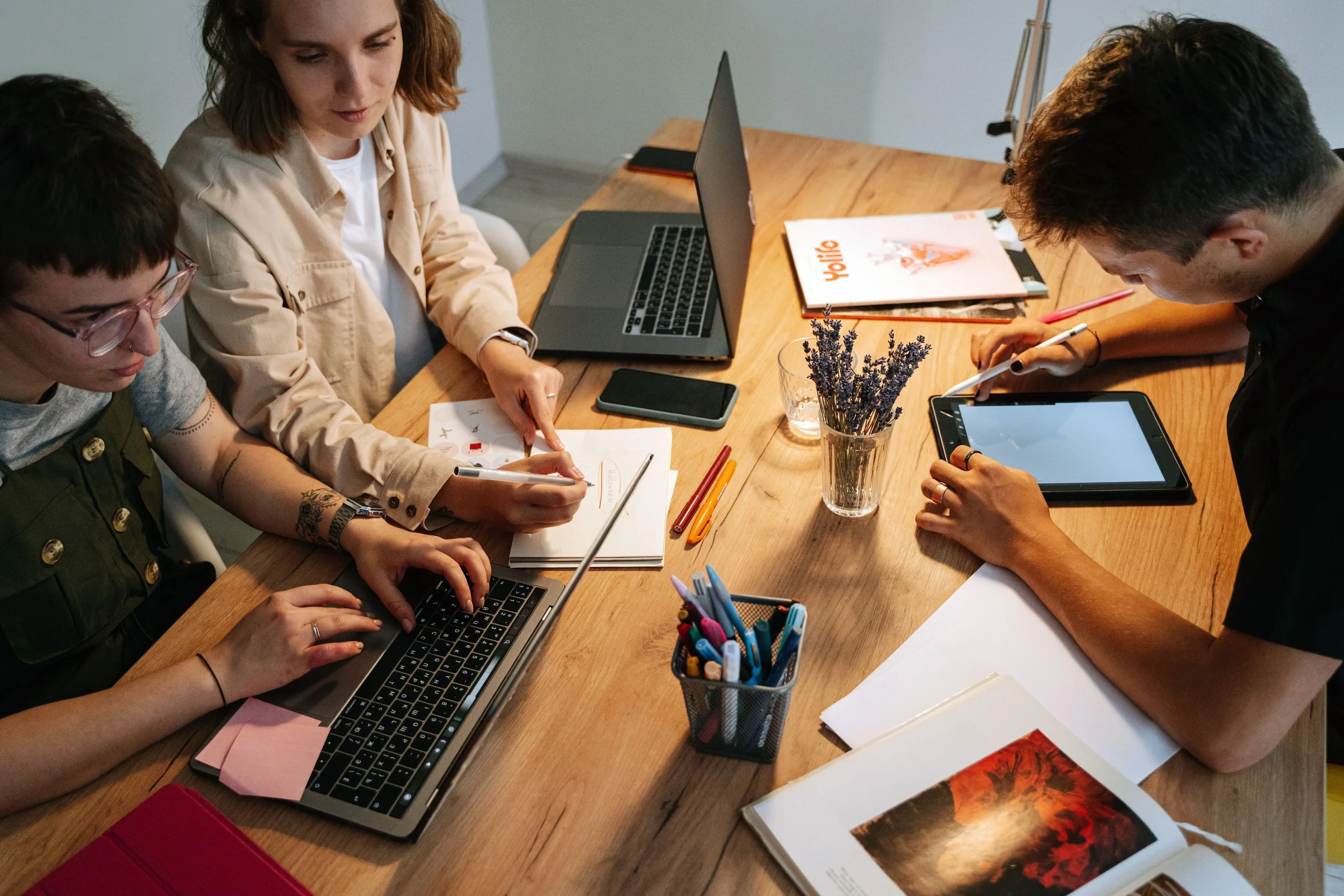 3 Studenten sitzen an einem Tisch. Alle haben einen Laptop vor sich und viele Dokumente sind auf dem Tisch.