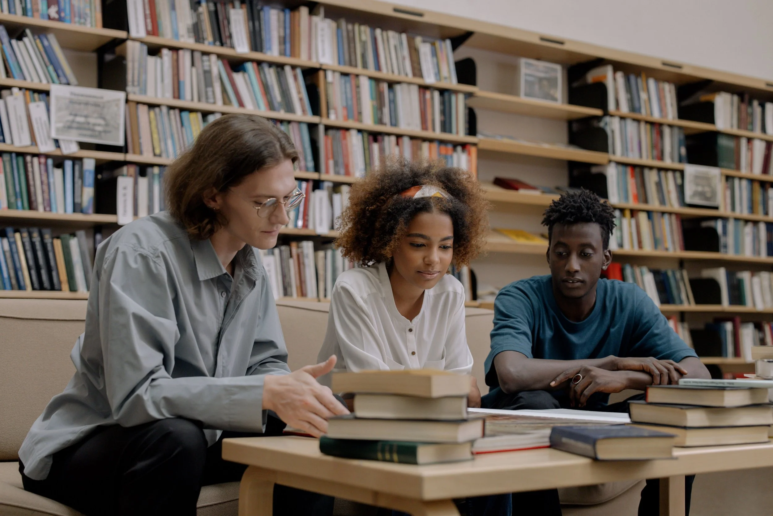 3 Studenten an einem Tisch in einer Bibliothek mit vielen Büchern auf dem Tisch.