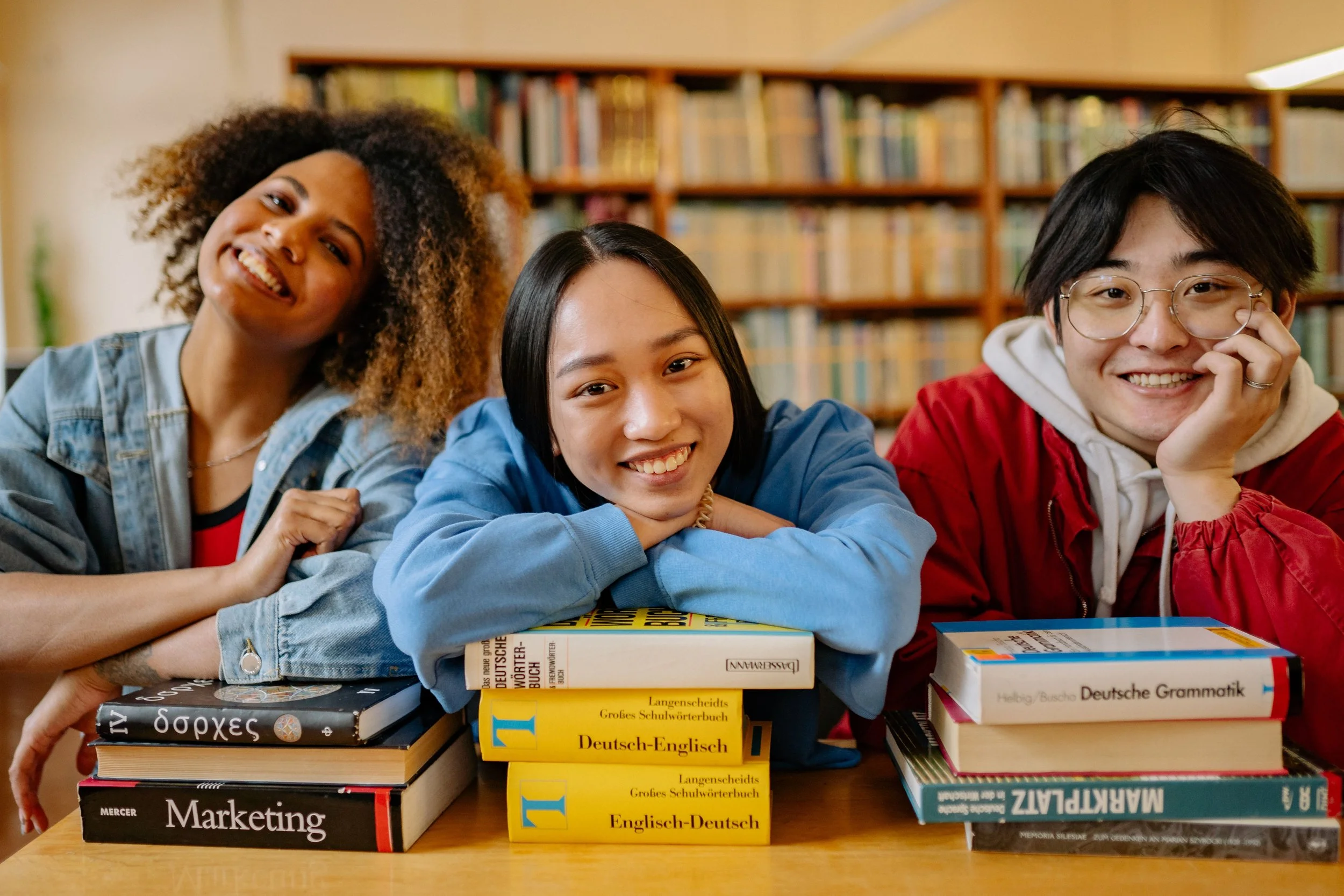 Drei junge Studenten lachen in die Kamera und stützen sich auf Deutschbüchern ab und lernen Deutsch in einer Bibliothek.