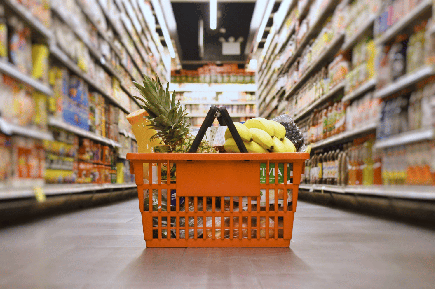 Orange shopping basket filled with fruits and groceries on store aisle floor, shelves stocked with various products in supermarket.