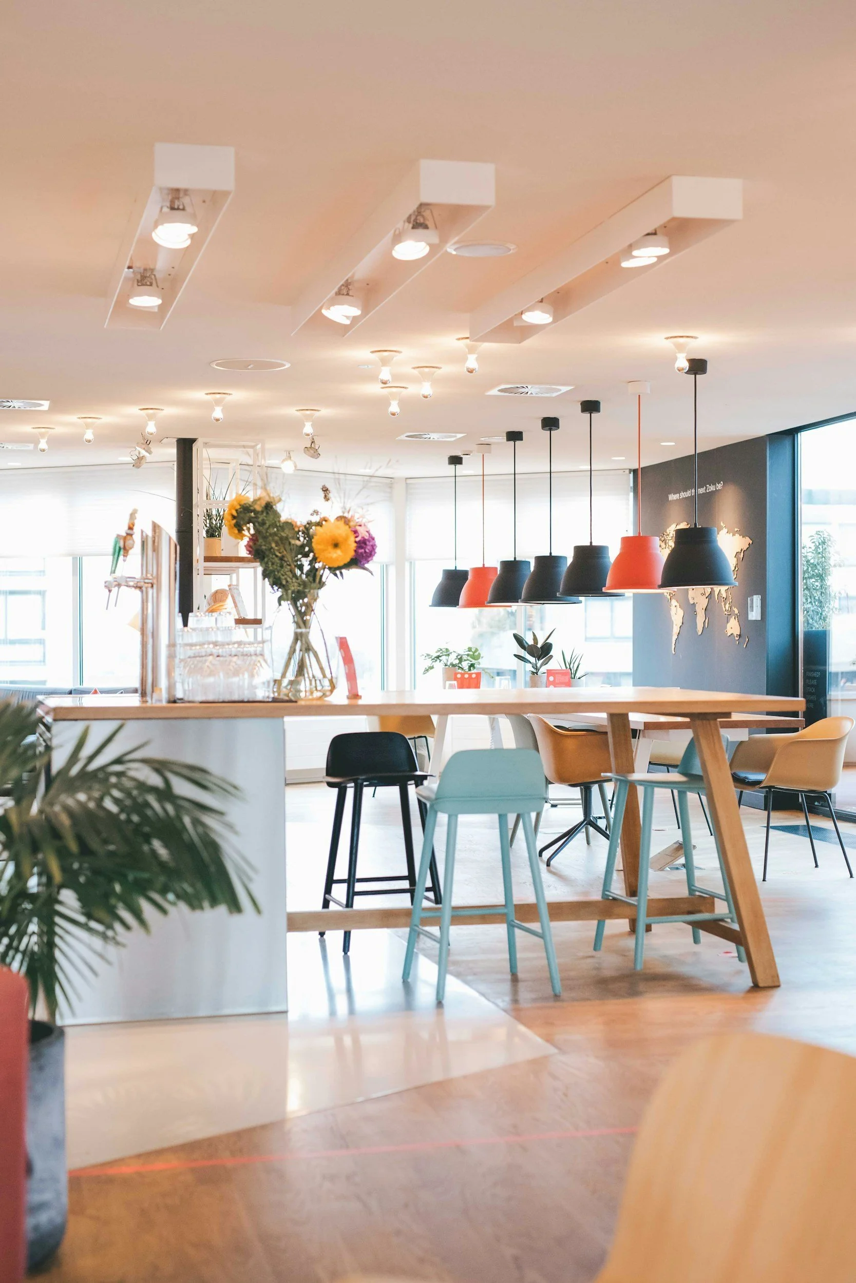 Interior of a modern cafe or restaurant with hanging pendant lights, a table with a flower vase, and various chairs around a wooden table. There are large windows letting in natural light and a map on the wall.