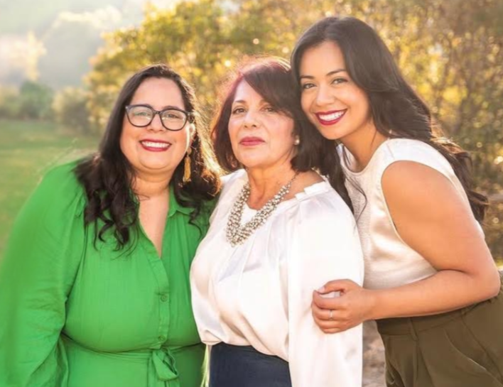 Three women smiling outdoors in a park, with trees and sunlight in the background, dressed stylishly.