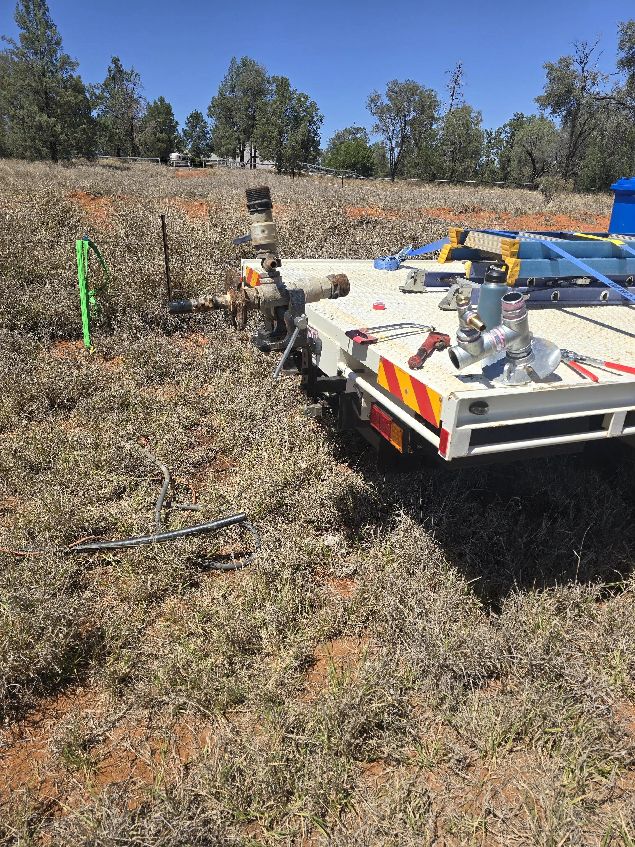 A utility truck with equipment and tools, including pipes, a water bottle, and a ladder, on the back, parked on a grassy field with dry grass and dirt, surrounded by trees and a clear blue sky.