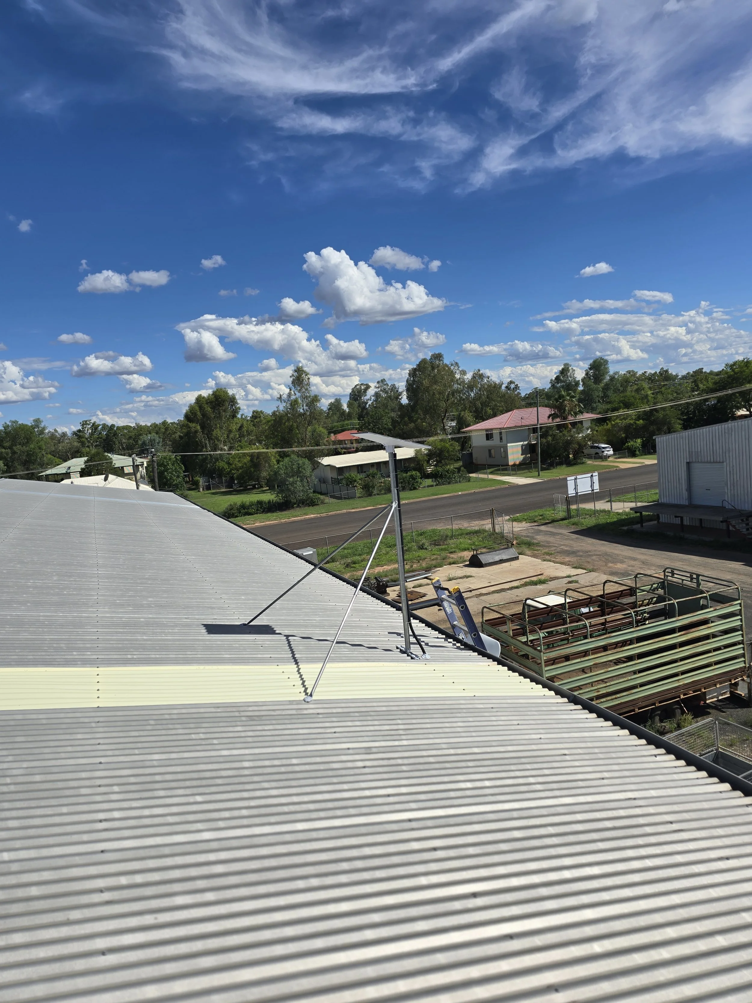 View of a suburban neighborhood with trees, houses, and a road, under a blue sky with scattered clouds, taken from a rooftop with solar panels and communication antennas.