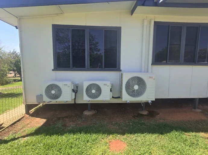 Three air conditioning units installed outside a house beneath two windows, with grass and dirt surrounding them.