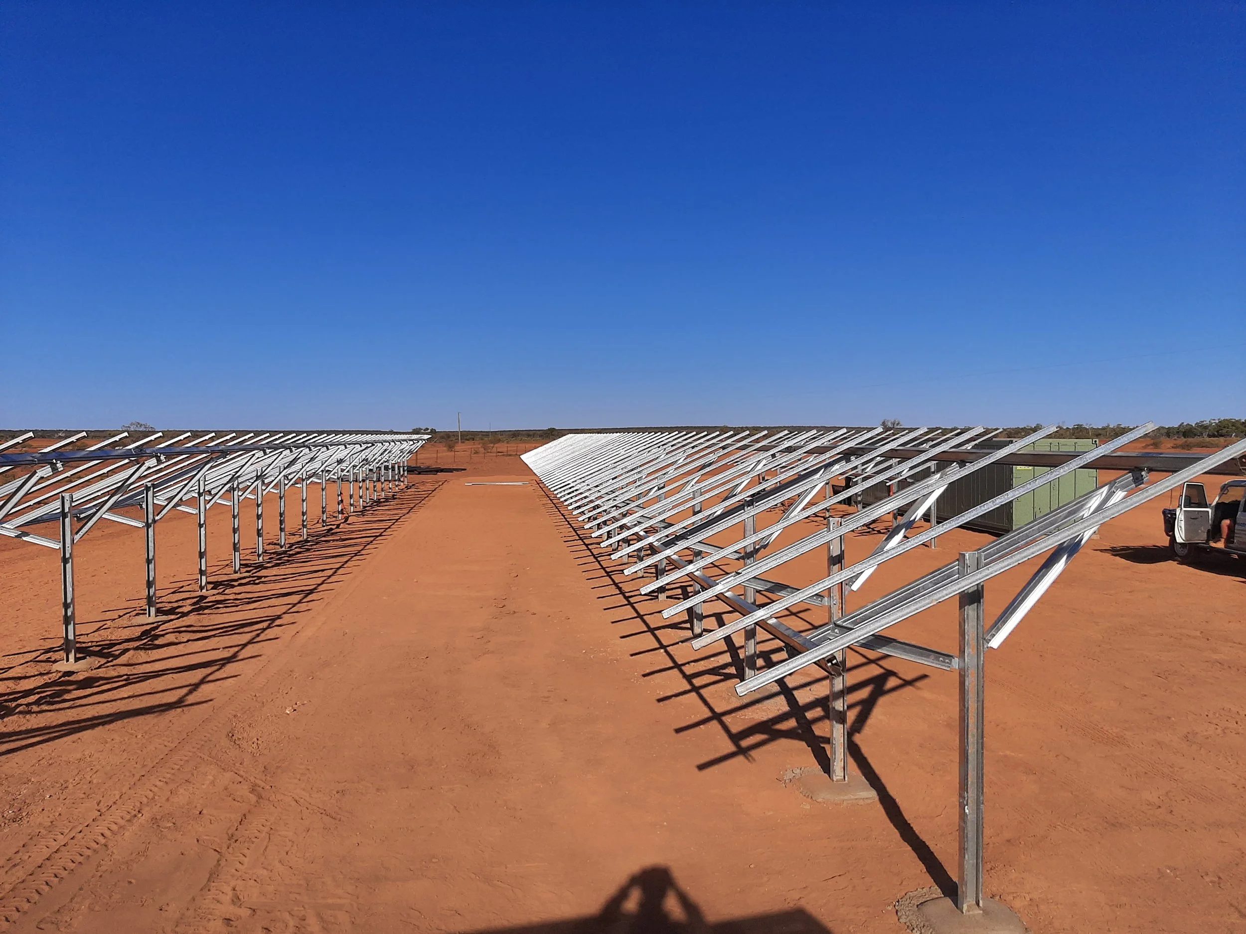 Solar panel mounts are being installed on a reddish dirt field under a clear blue sky, with a few trees in the distance.