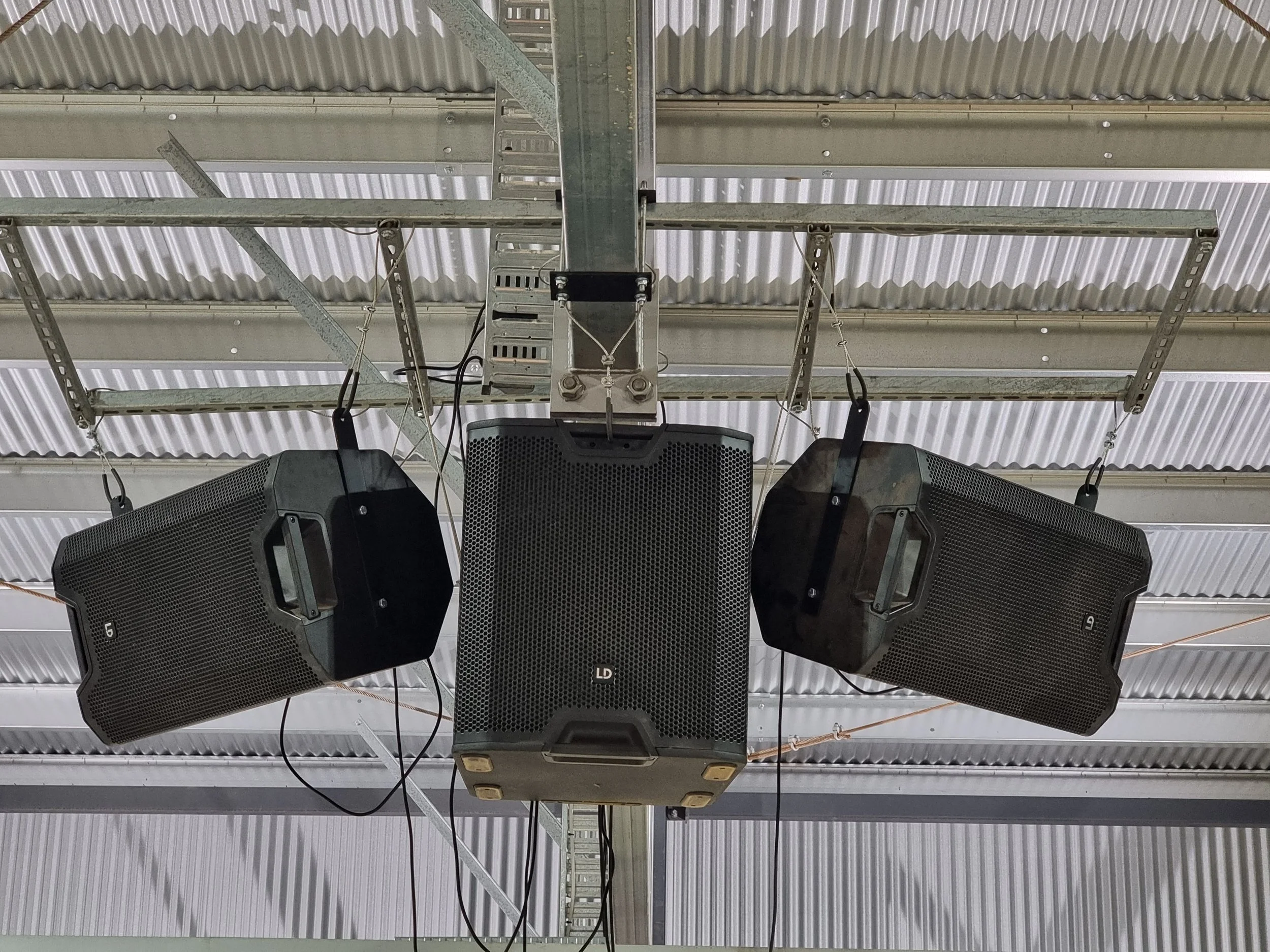 Four large black speakers hanging from the ceiling of an industrial or warehouse building.