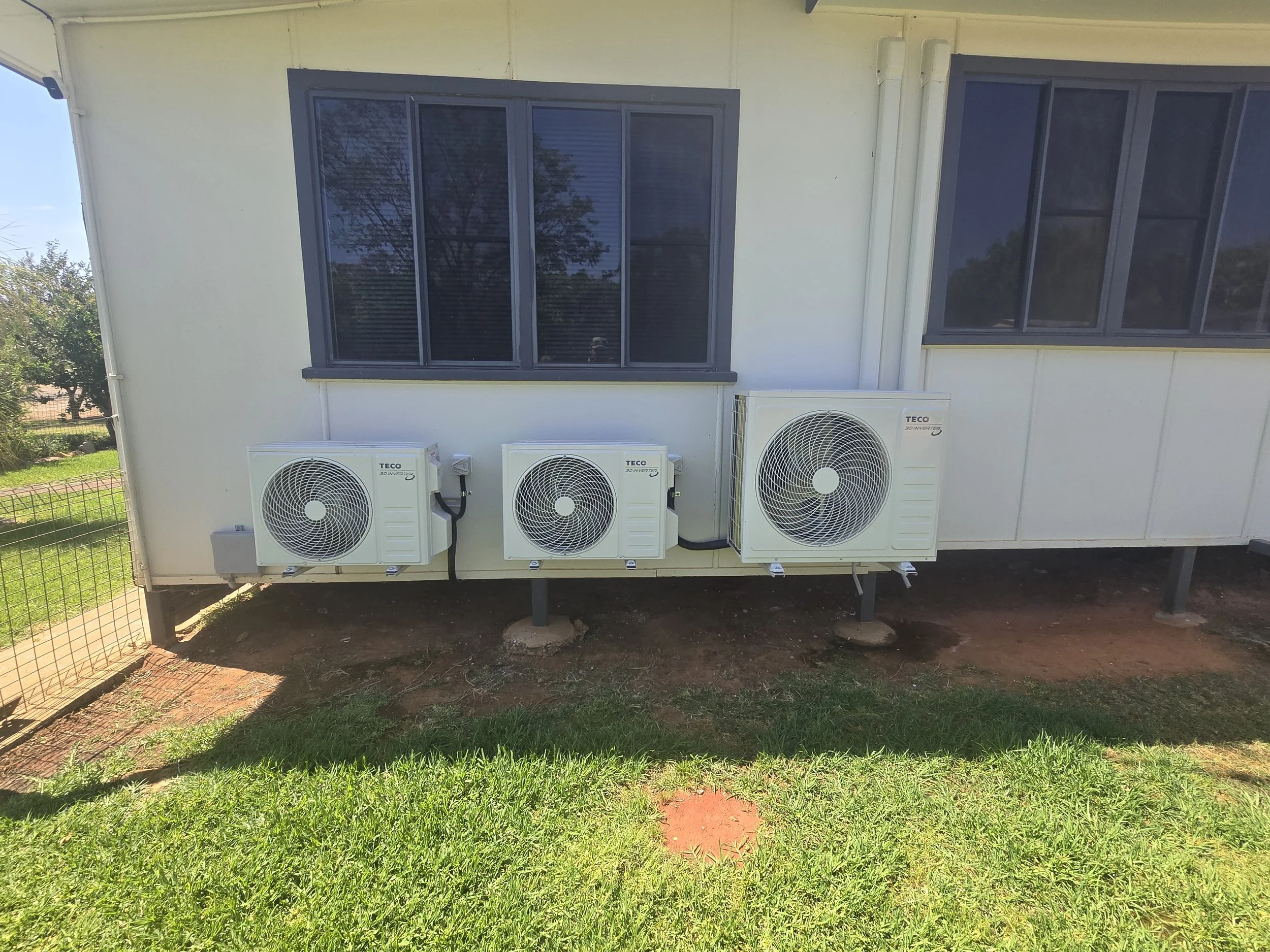Exterior of a house with three air conditioning units mounted on the wall, a large window with dark frames, and a small grassy yard in front.
