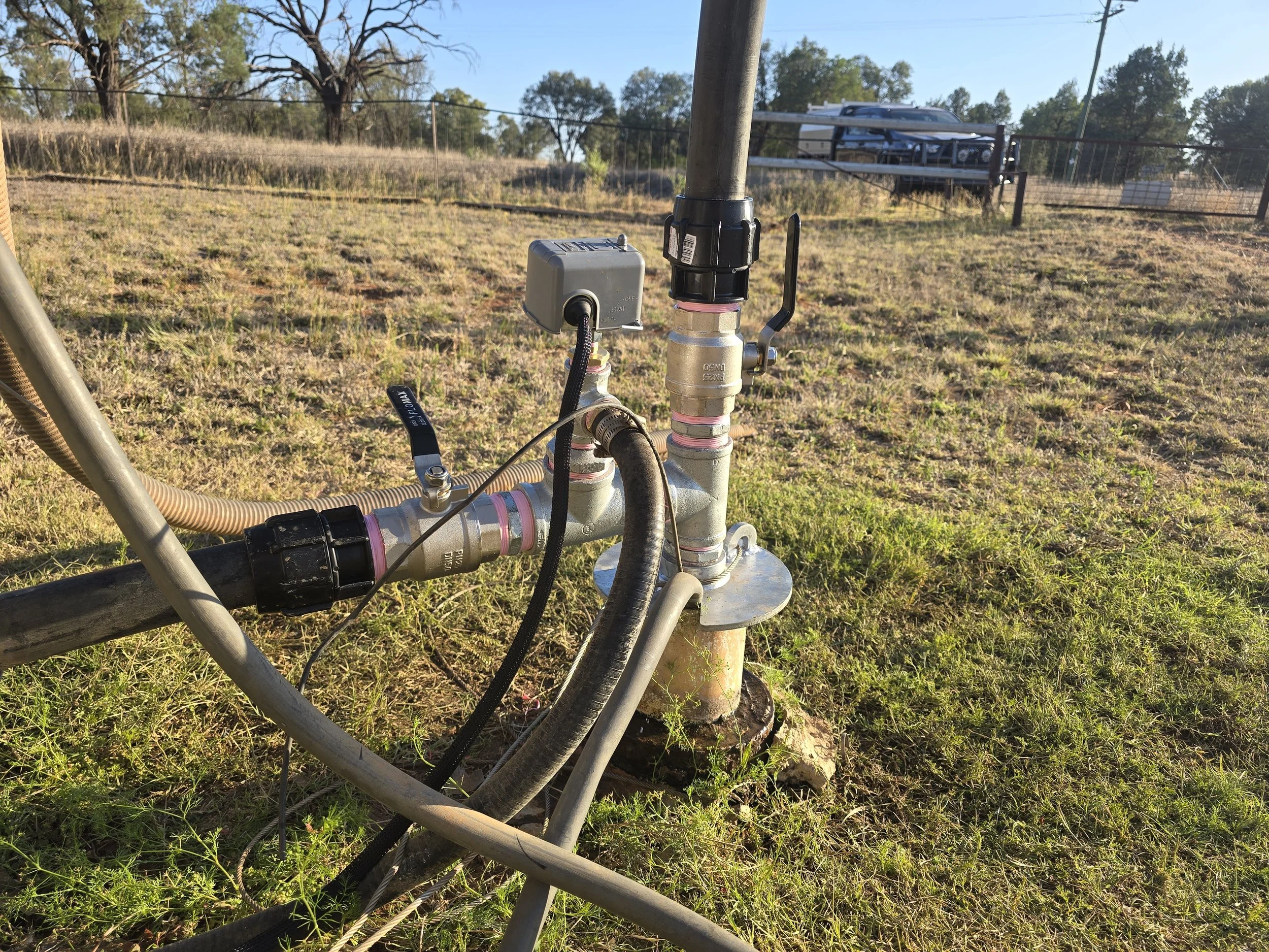 Close-up of a water pump system with pipes, valves, and an electrical box, set outdoors on grassy ground with trees in the background.