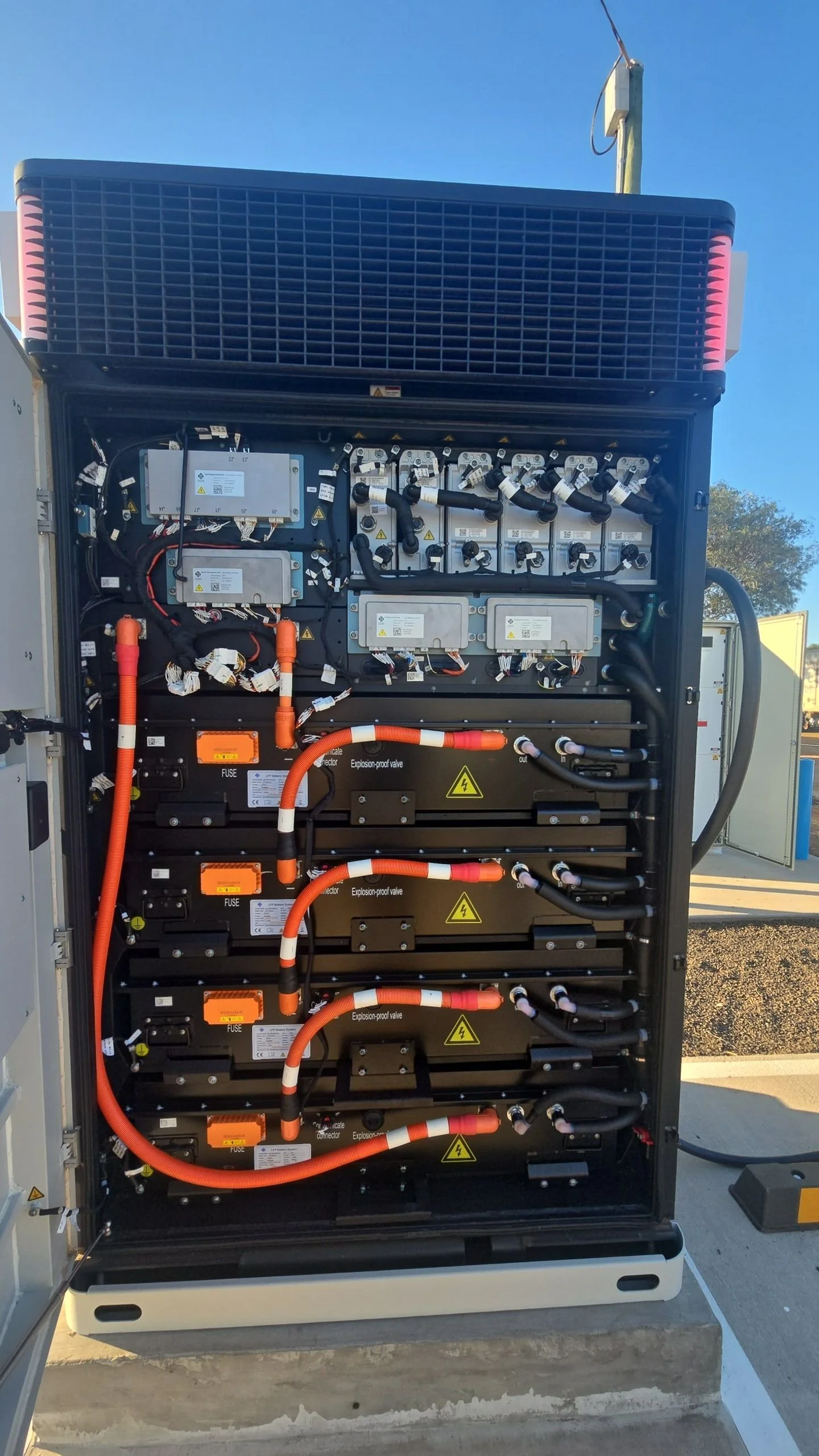 Open electrical cabinet with various wires, circuit boards, and hazard labels, outdoors against a clear blue sky.