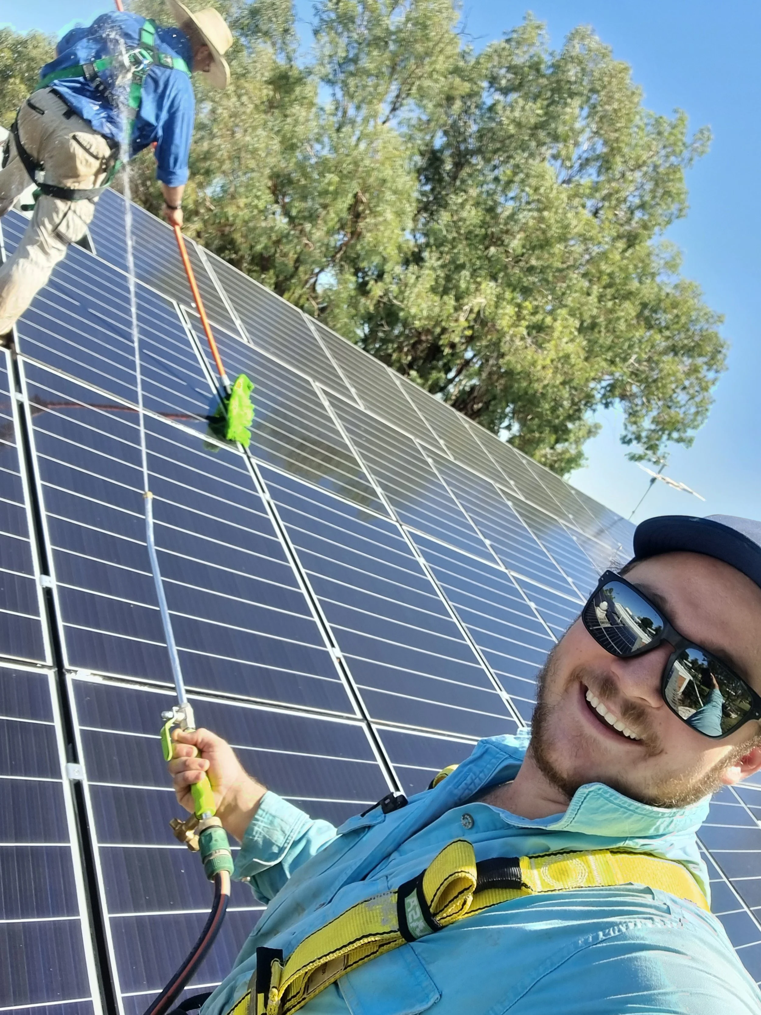 A man with a beard and sunglasses smiling, wearing a blue shirt with yellow suspenders, working on solar panels with a hose, outdoors with trees and a clear blue sky in the background.