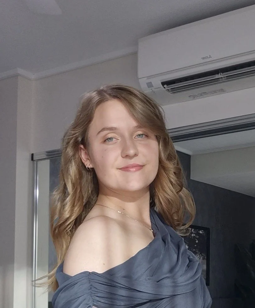 A young woman with light brown hair and blue eyes, wearing a dark off-shoulder dress, smiling softly inside a room with an air conditioner on the wall behind her.