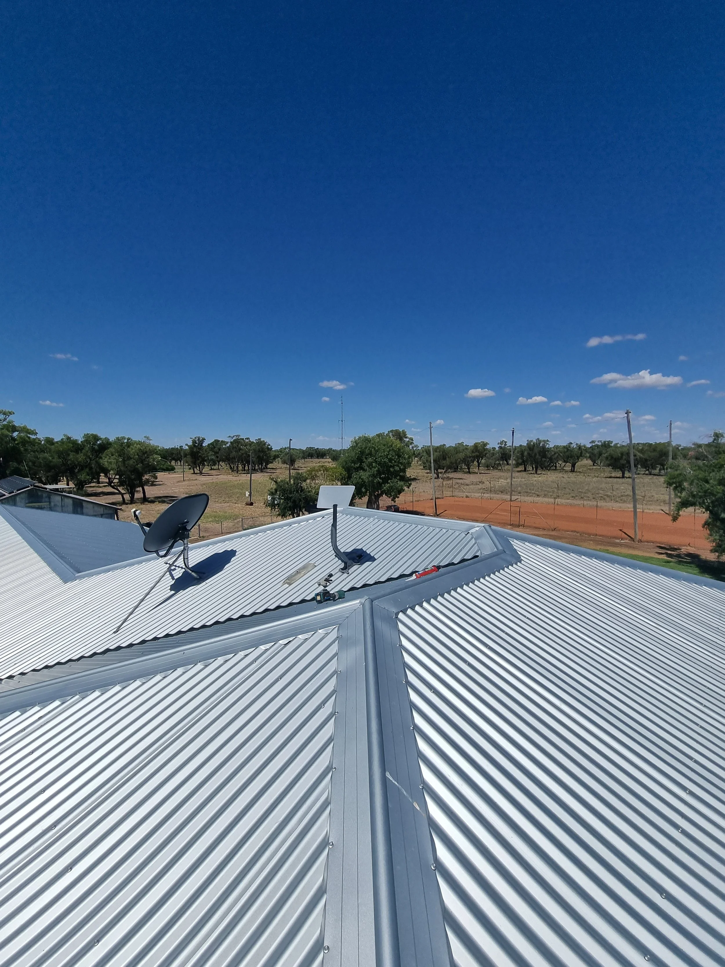 Metal roof with two satellite dishes, and a background of trees, dirt area, power lines, and a blue sky with some scattered clouds.