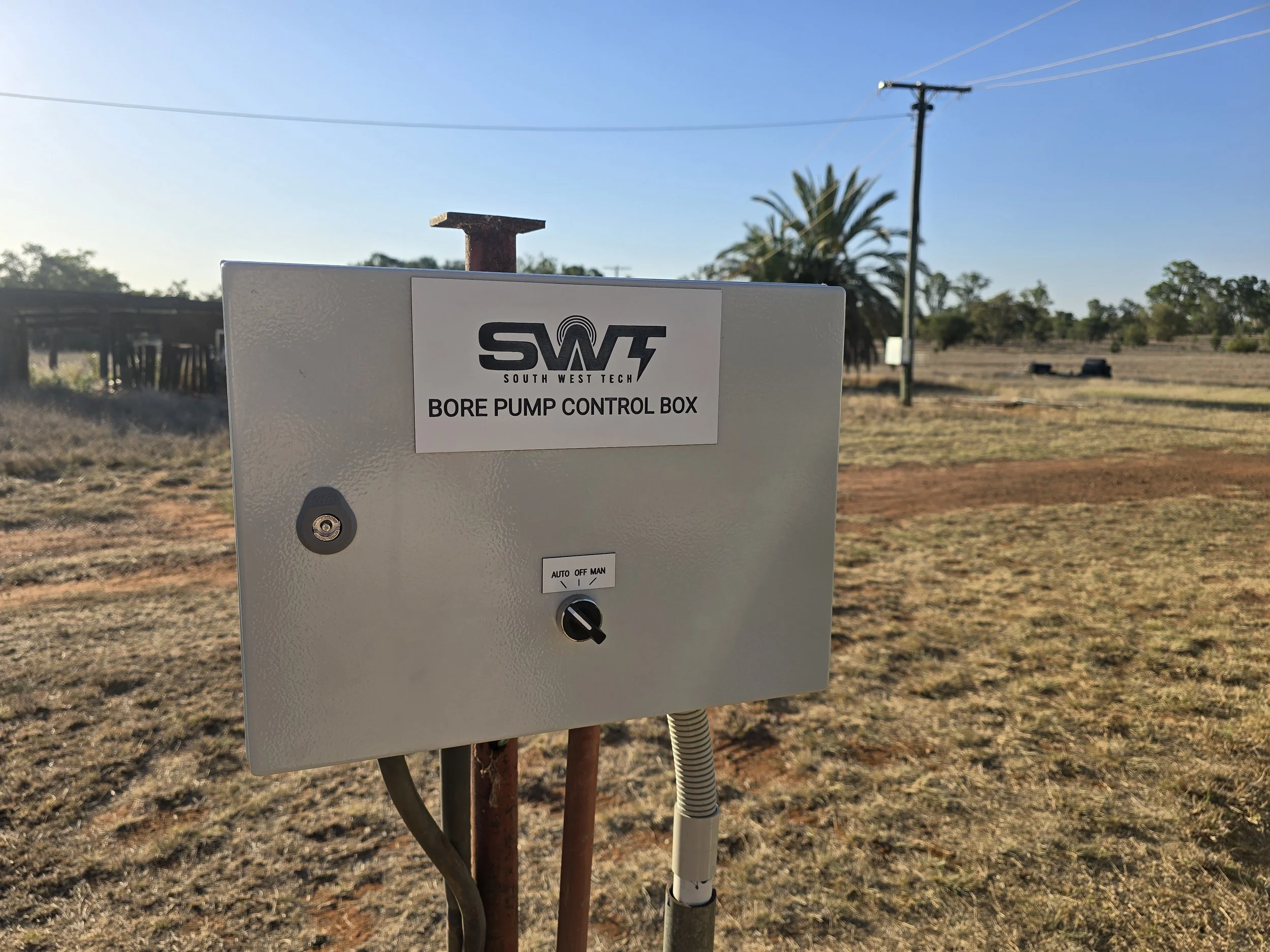 Electrical control box labeled 'South West Tech. Bore Pump Control Box' outdoors in a rural area with trees, power lines, and a clear blue sky in the background.