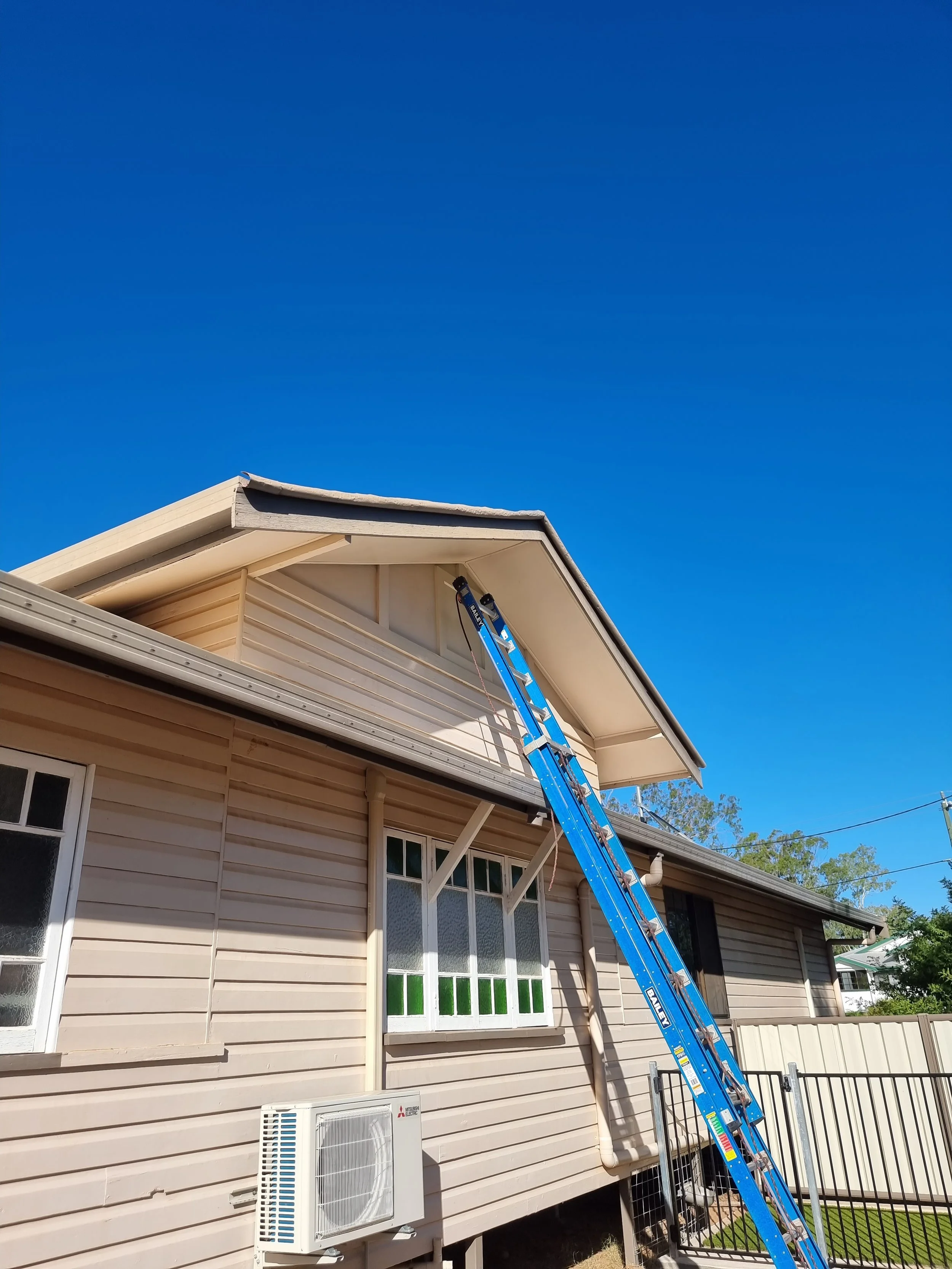 A house with beige siding, a gabled roof, and a large window with green and white panes. There is a blue ladder leaning against the house, reaching the roof. A white air conditioning unit is installed on the exterior wall.