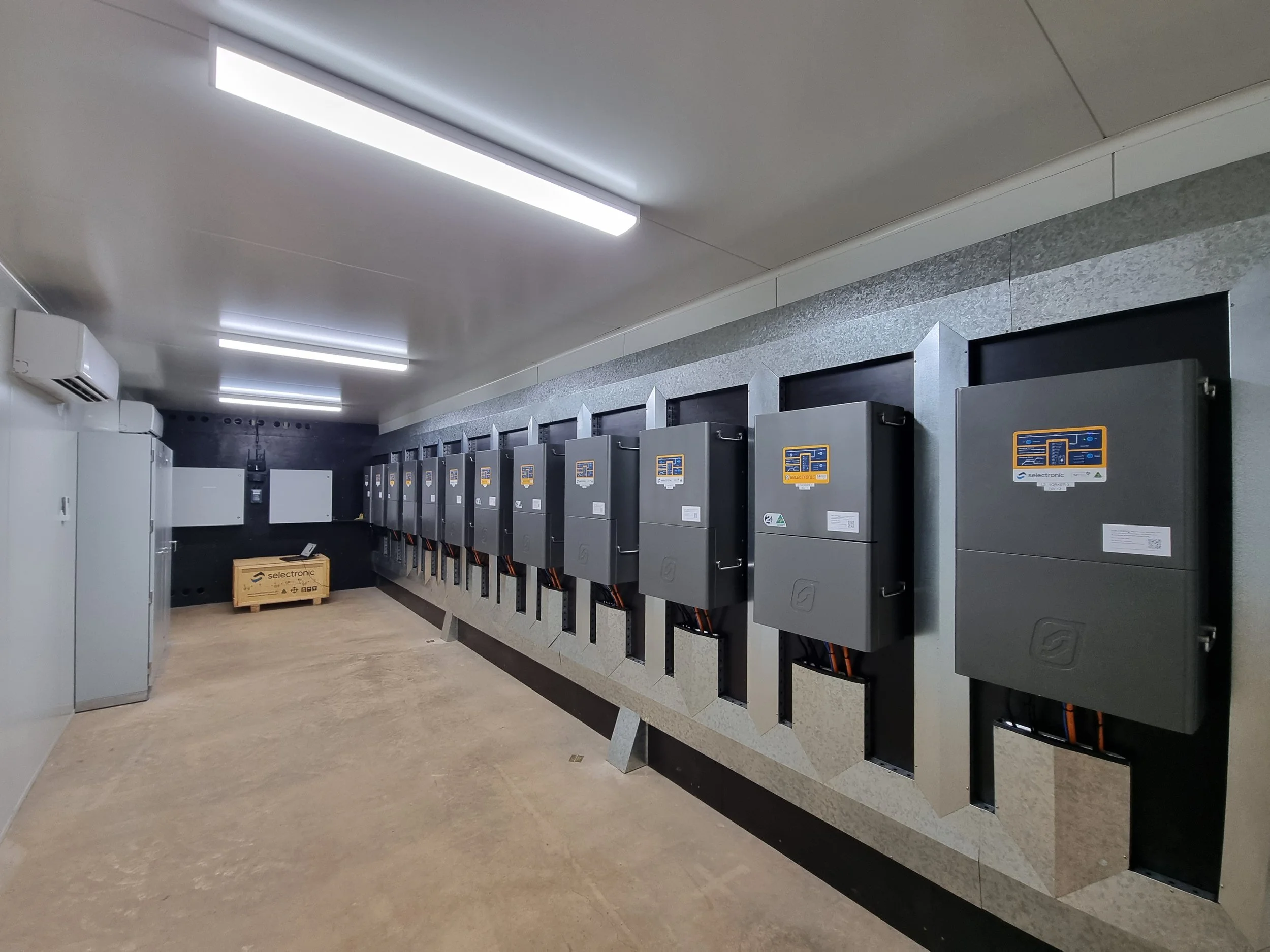 An indoor room with a row of black electrical or control panels mounted on a wall, a wooden crate on the floor, and an air conditioning unit on the wall. The ceiling has rectangular fluorescent lights.