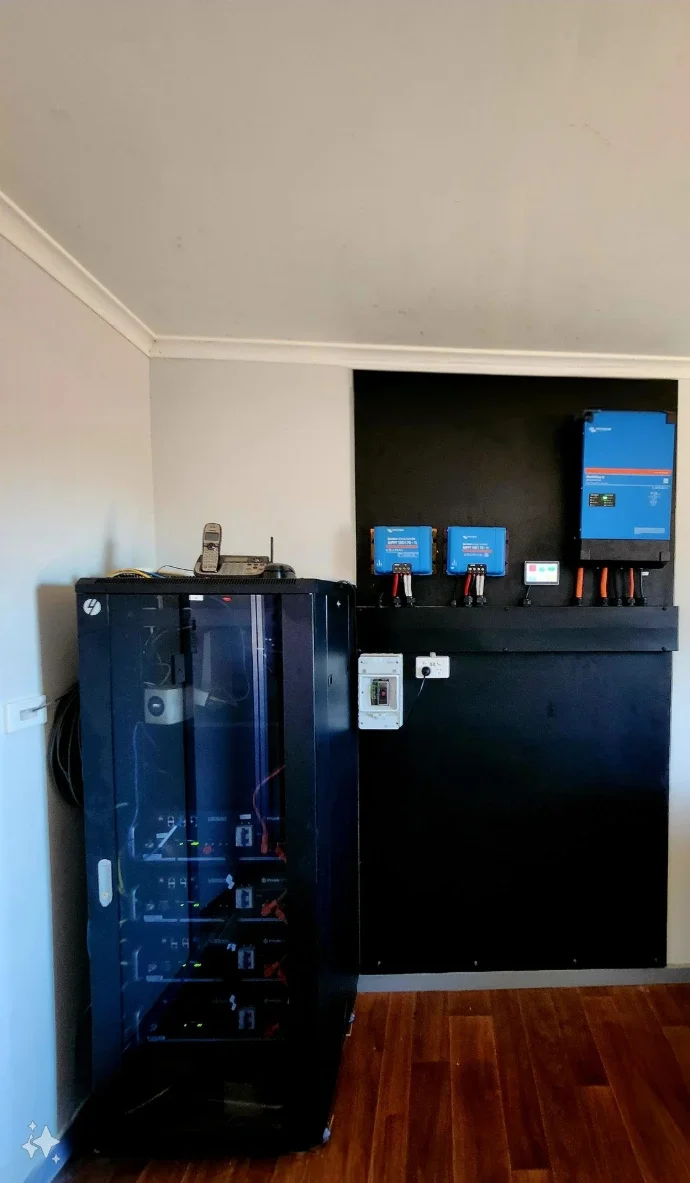 A black server rack with multiple electronic devices inside, next to a black control panel with blue devices mounted on it, on a wooden floor in a room with beige walls.