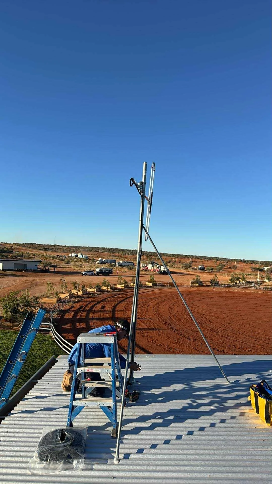 Person installing an antenna on a rooftop with a rural landscape, dirt racetrack, and parked vehicles in the background on a sunny day.