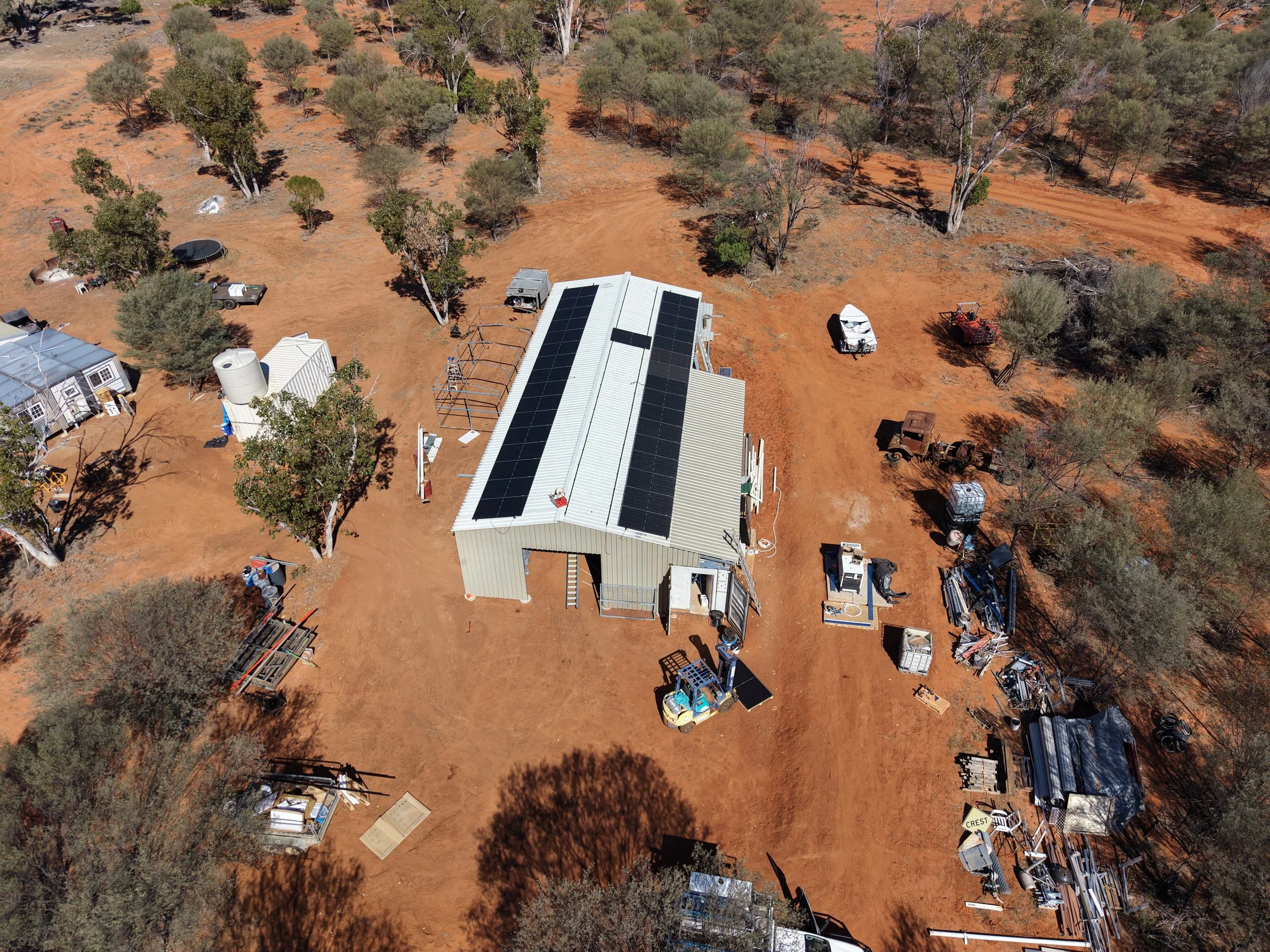 Aerial view of a rural property with trees, dirt roads, and various equipment and vehicles, including solar panels on a building roof.