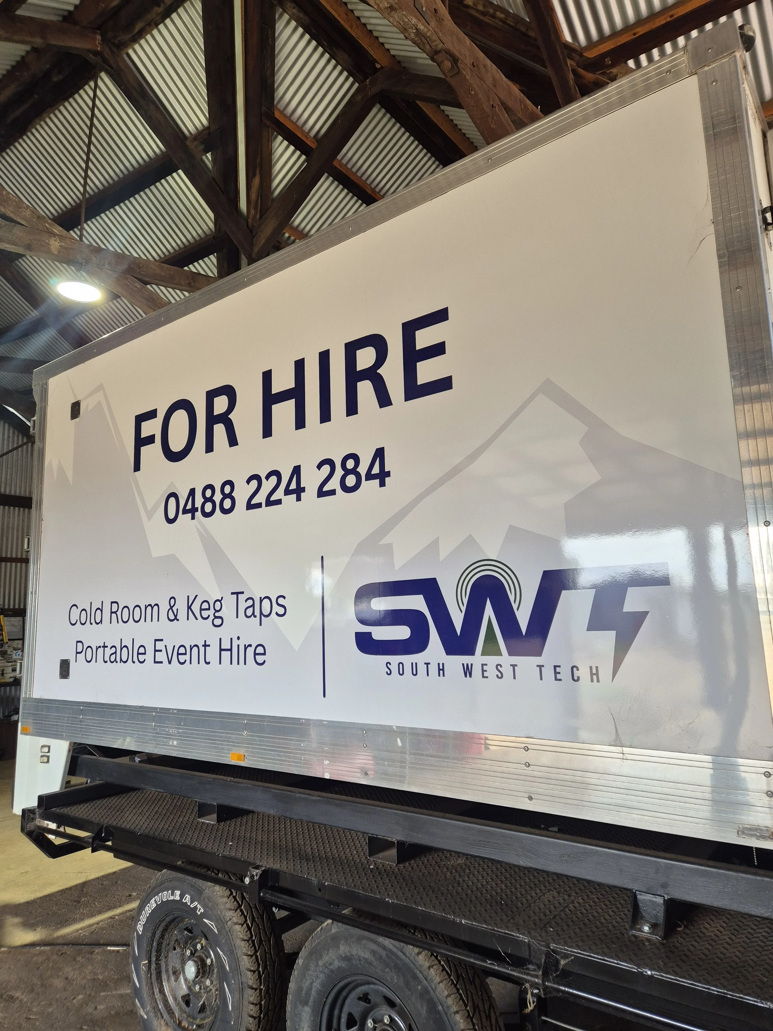 A large white trailer with signage for South West Tech is parked inside a building with a metal roof and wooden beams. The sign indicates services such as event hire, cold room, keg taps, and portable event hire, along with a phone number.