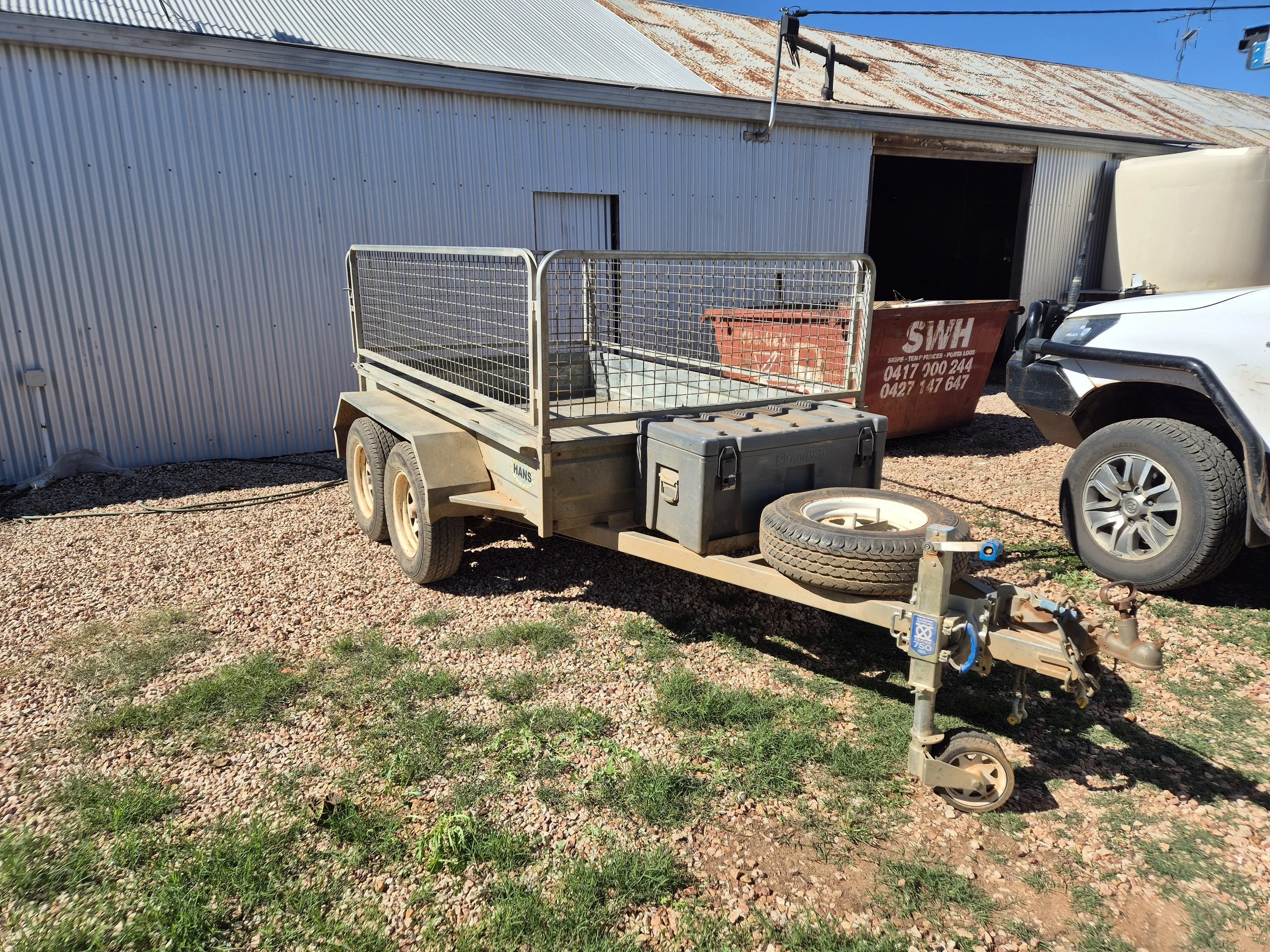 An outdoor scene featuring a corrugated metal building with a rusty roof, a white utility vehicle, a trailer with a metal cage, a red dumpster, a spare tire, and various tools and equipment on the ground.