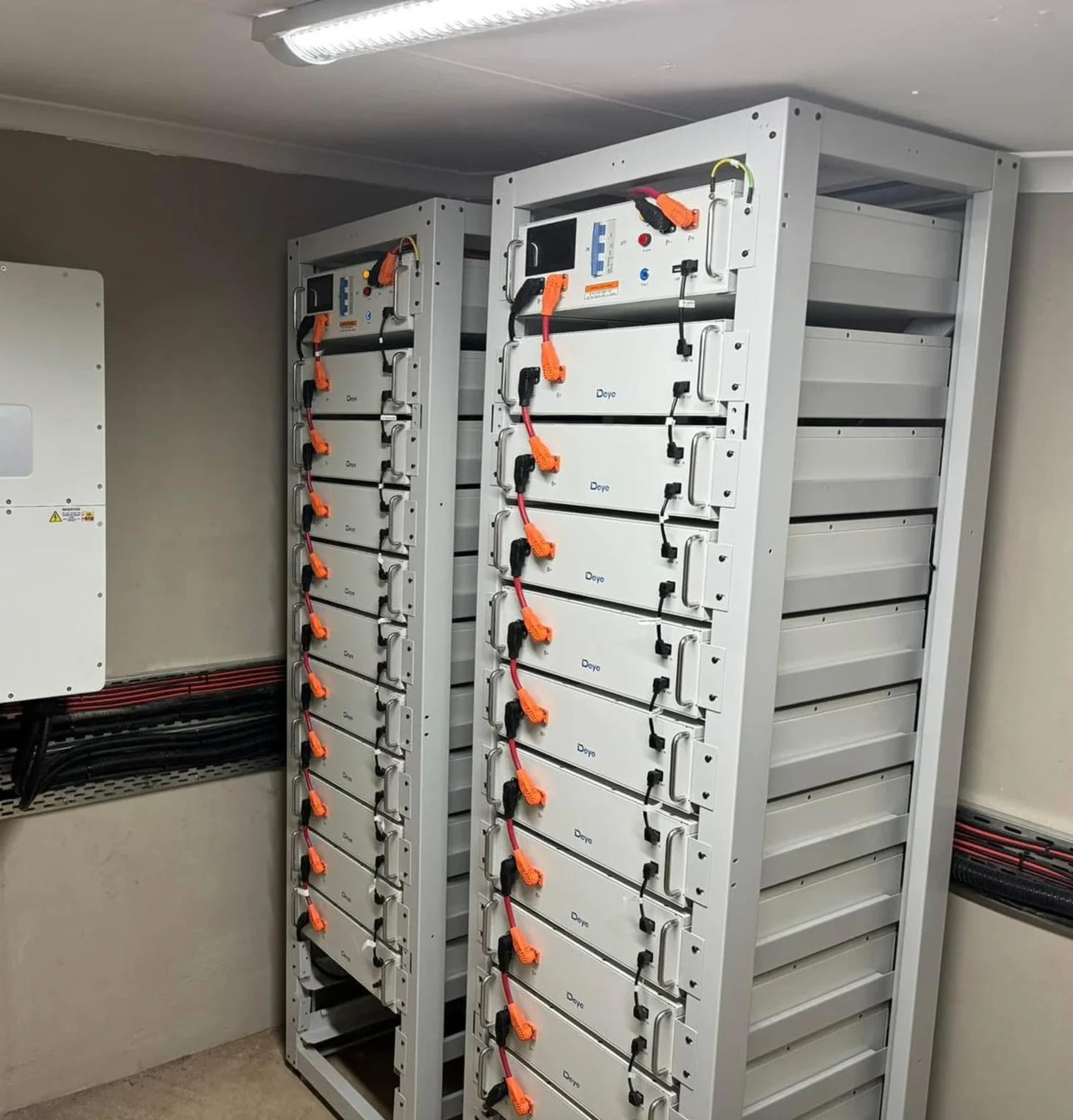 Two tall, white server racks with orange and black cables connected to the front, inside a room with a gray wall and ceiling-mounted light.