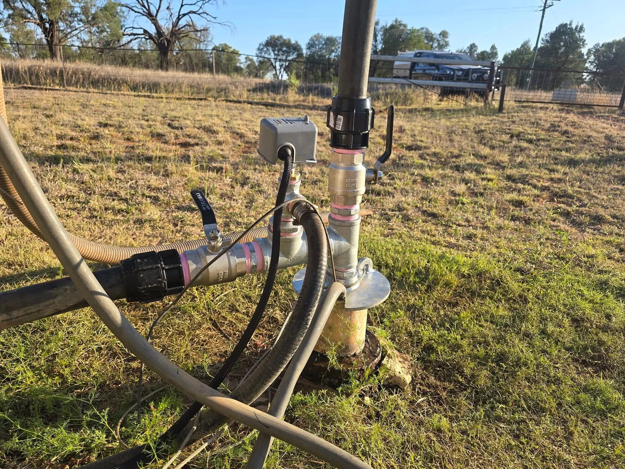 Close-up of underground water piping with a valve, fittings, and cables in an outdoor grassy area, with a fenced background and trees.
