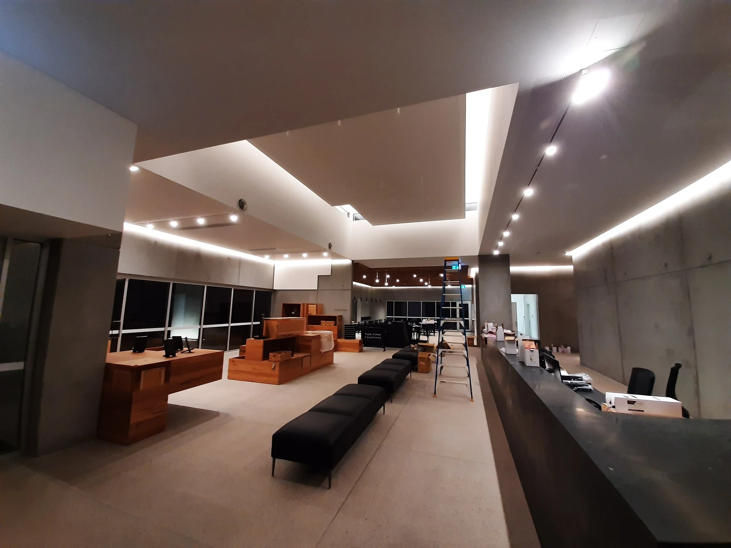 A modern, well-lit interior space with high ceilings, concrete walls, and a series of black benches arranged along a curved wall. There are wooden furniture pieces and a ladder in the background, possibly indicating ongoing setup or renovation.