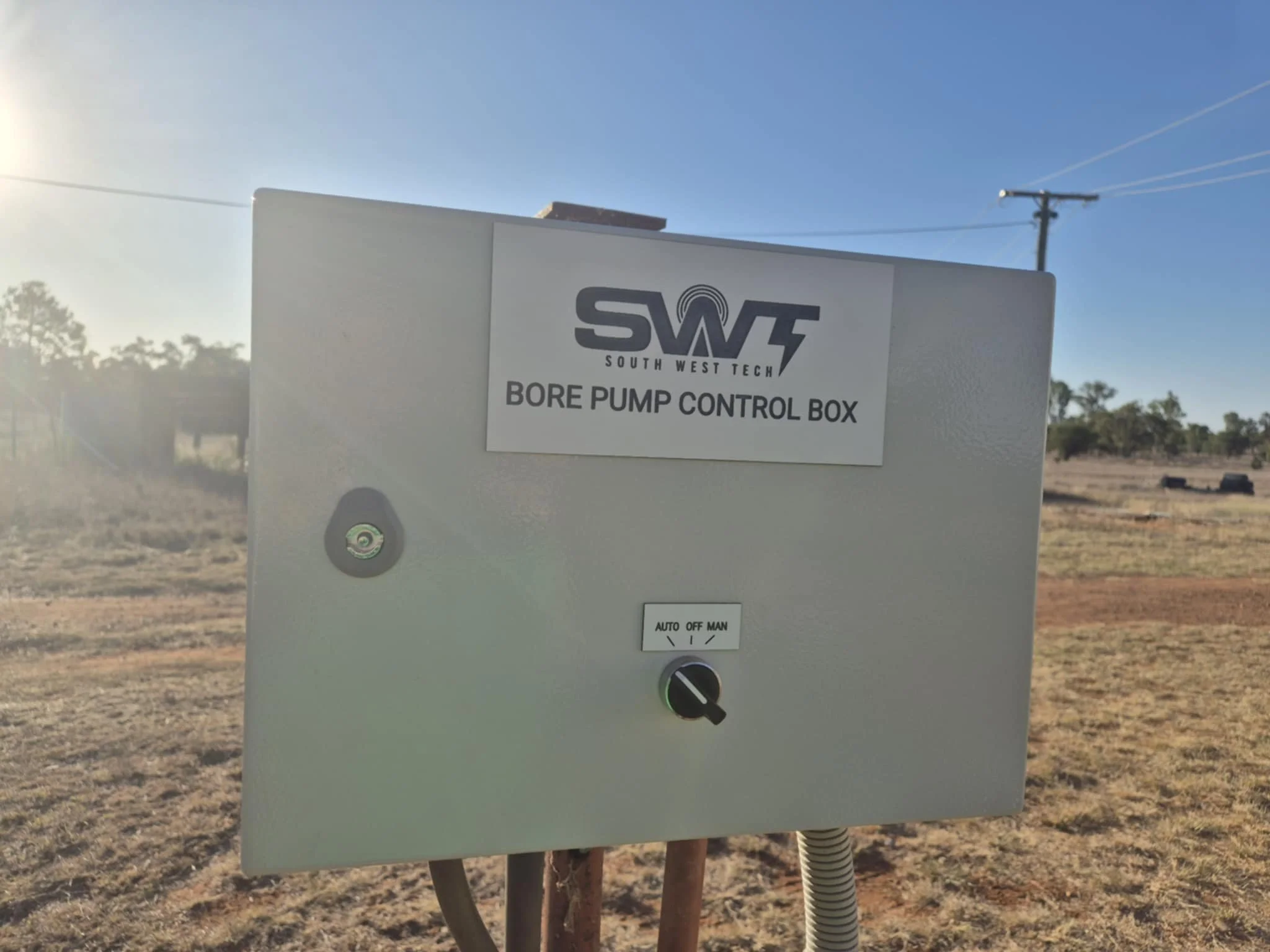 Close-up of a gray control box labeled "South West Tech Bore Pump Control Box" with a small lock and an on/off switch, set outdoors against a background of a rural landscape with trees and a clear sky.
