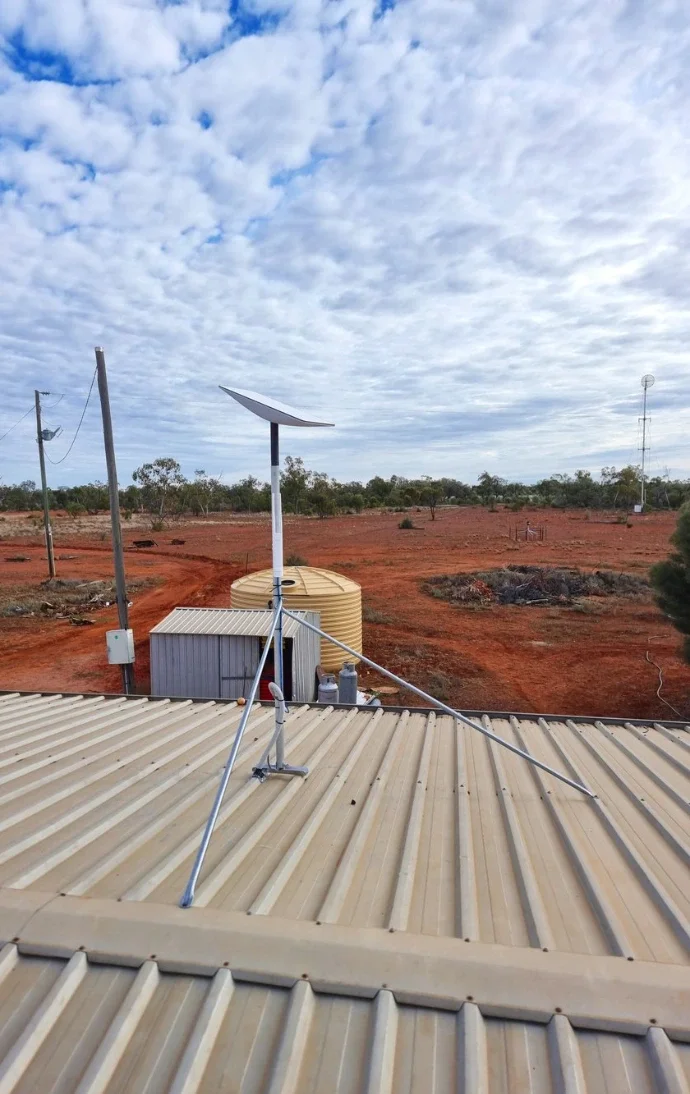 View from a rooftop showing a rooftop mounted solar panel and antenna, desert terrain with sparse trees, electrical poles, a water tank, and a distant fire pit under a cloudy sky.