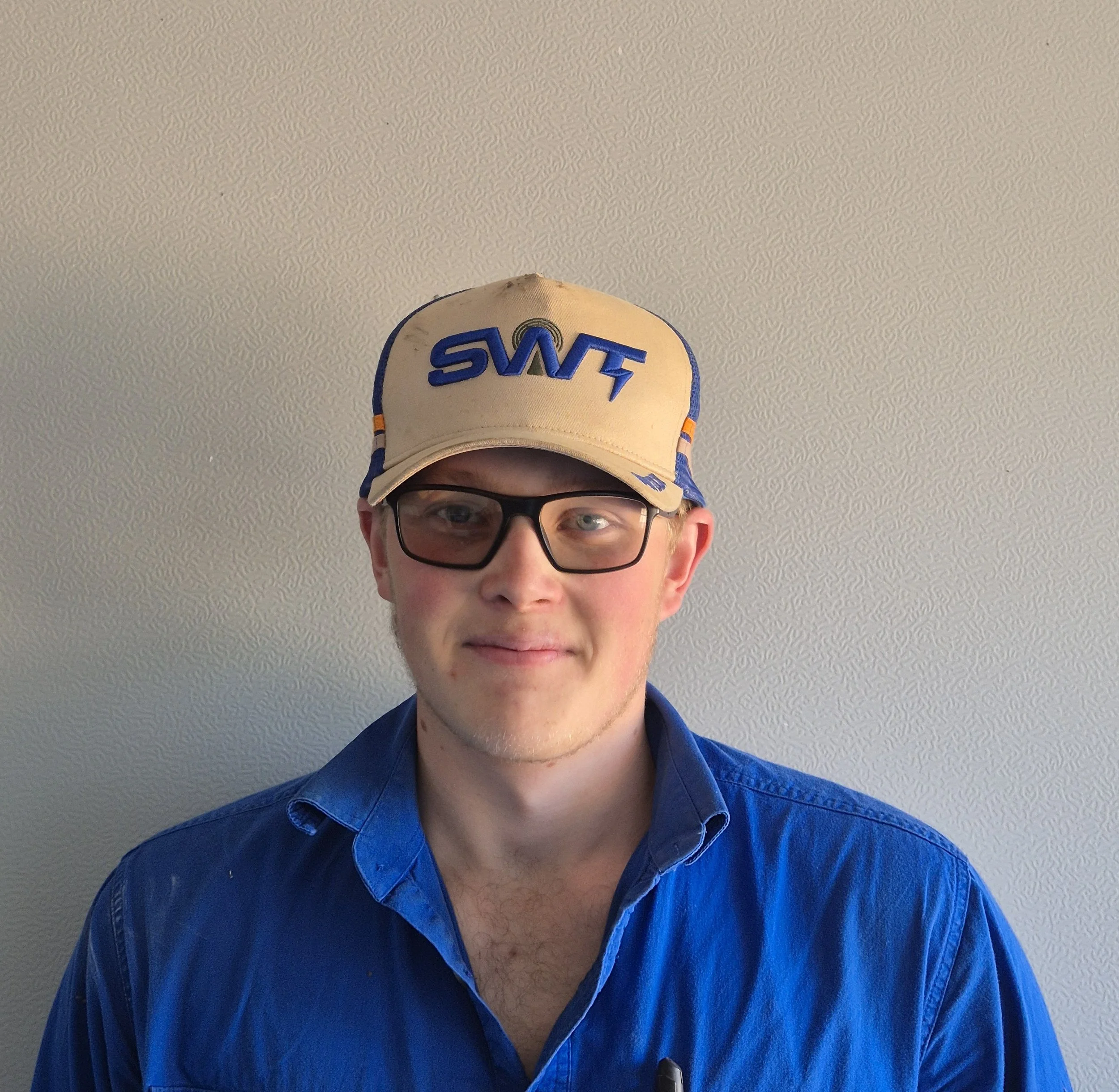 A person wearing glasses and a beige baseball cap with "SNT" embroidered on it. The person is wearing a blue shirt and the background is a plain, textured ceiling.