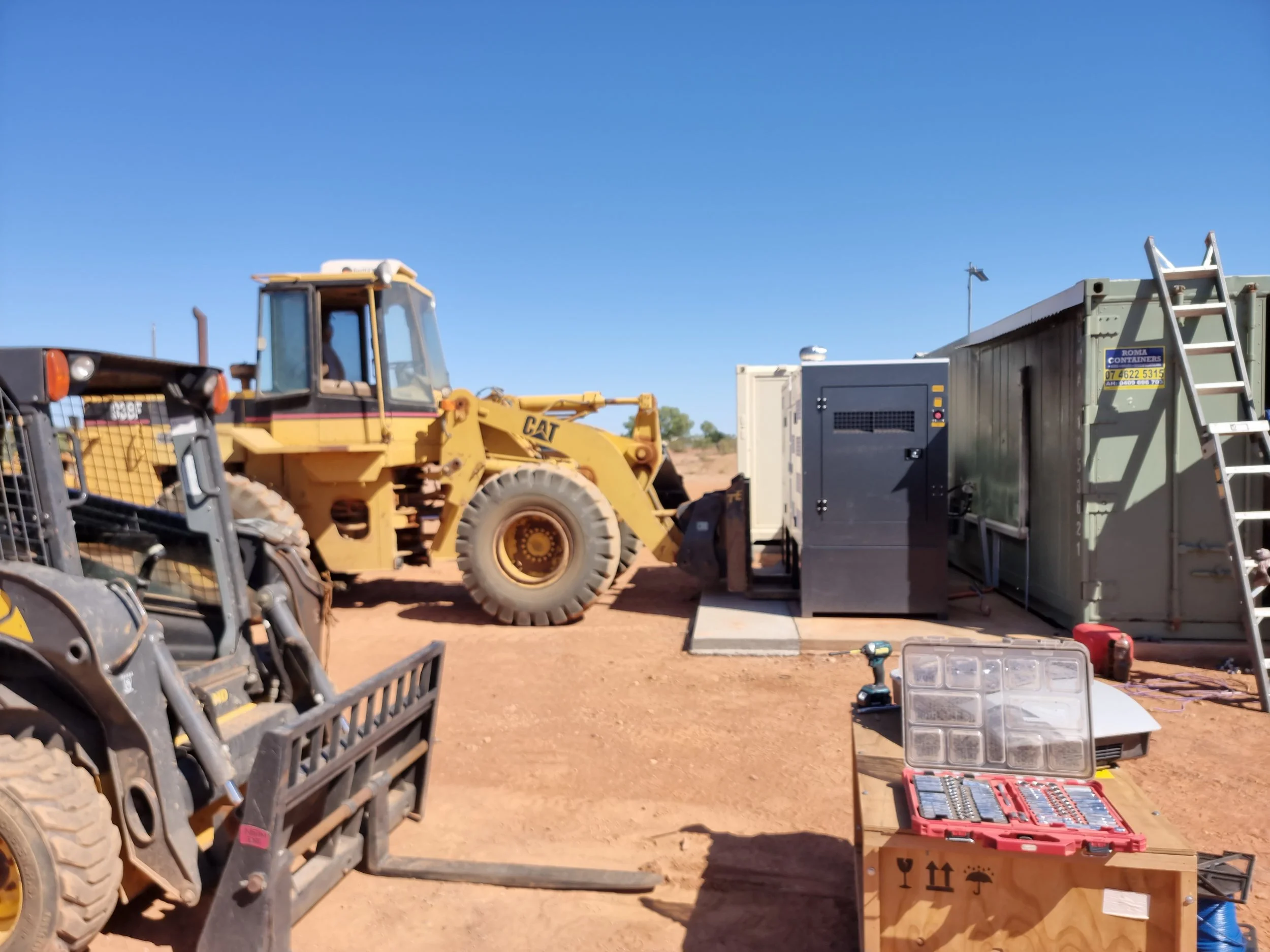 Various construction tools and equipment, including a yellow Caterpillar bulldozer, tents, a ladder, and toolboxes, set up outdoors under a clear blue sky.