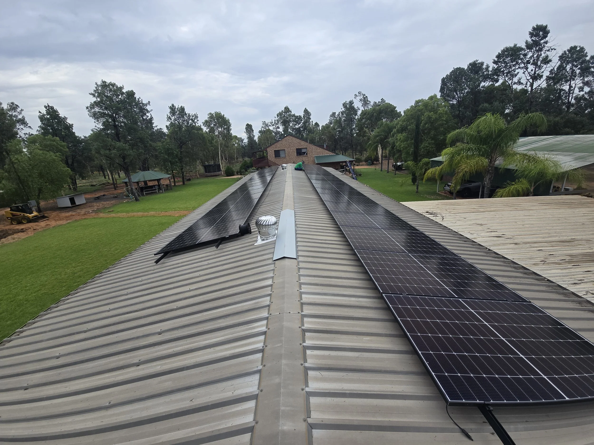 A residential roof with solar panels installed, surrounded by a green yard, trees, and a cloudy sky.