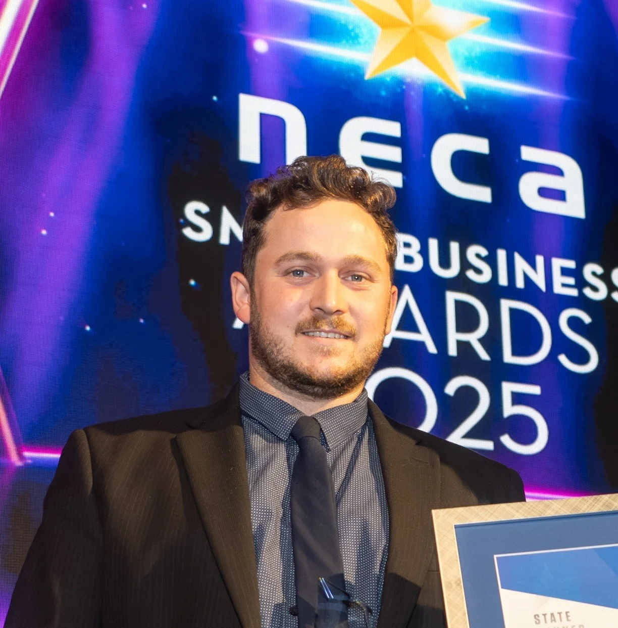 A man in a suit holding an award at the 2025 Nevada Small Business Awards ceremony, with a digital display in the background.