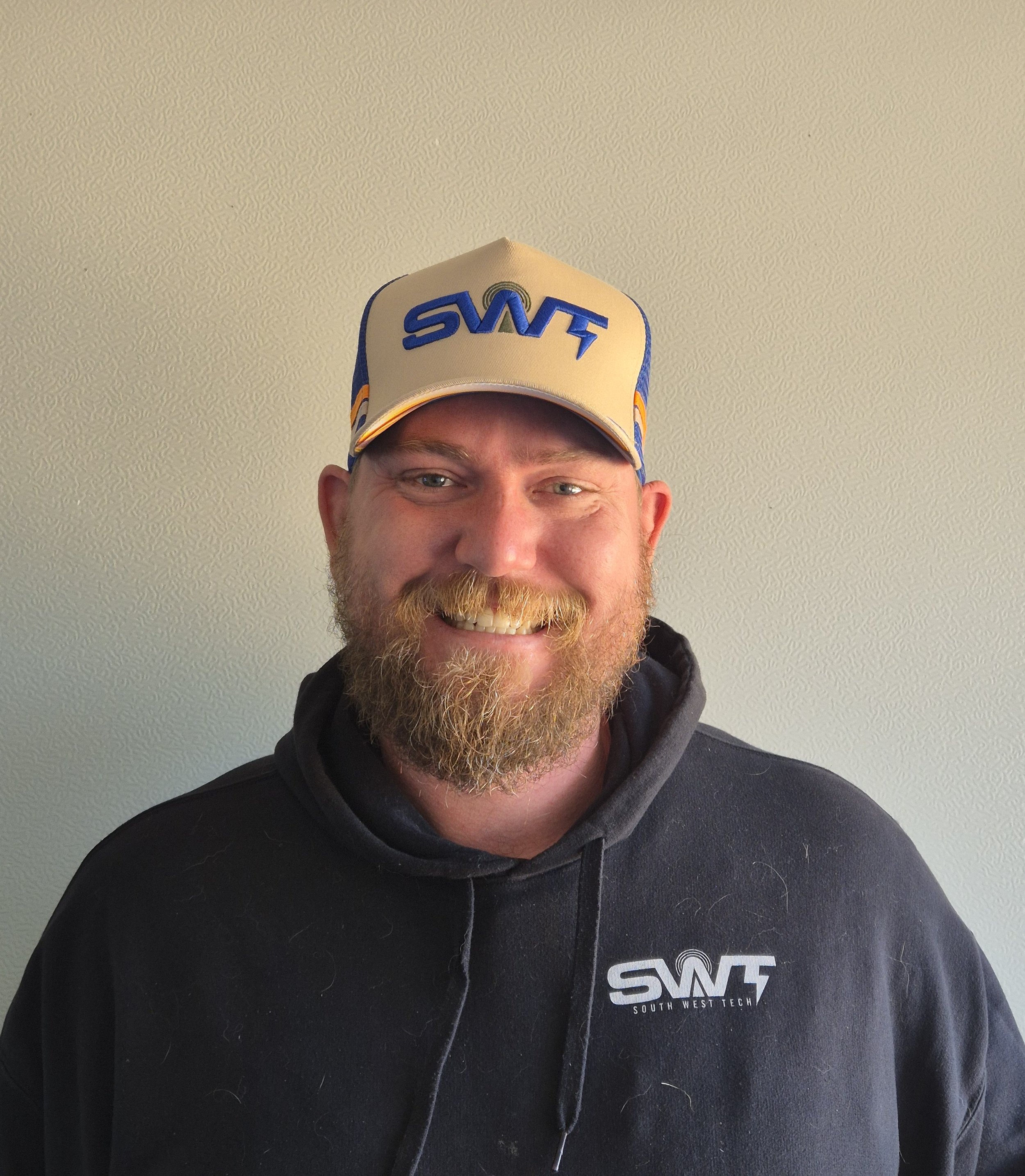 A smiling man with a beard and mustache wearing a black hoodie and a beige baseball cap with blue and orange accents, and the logo 'SW