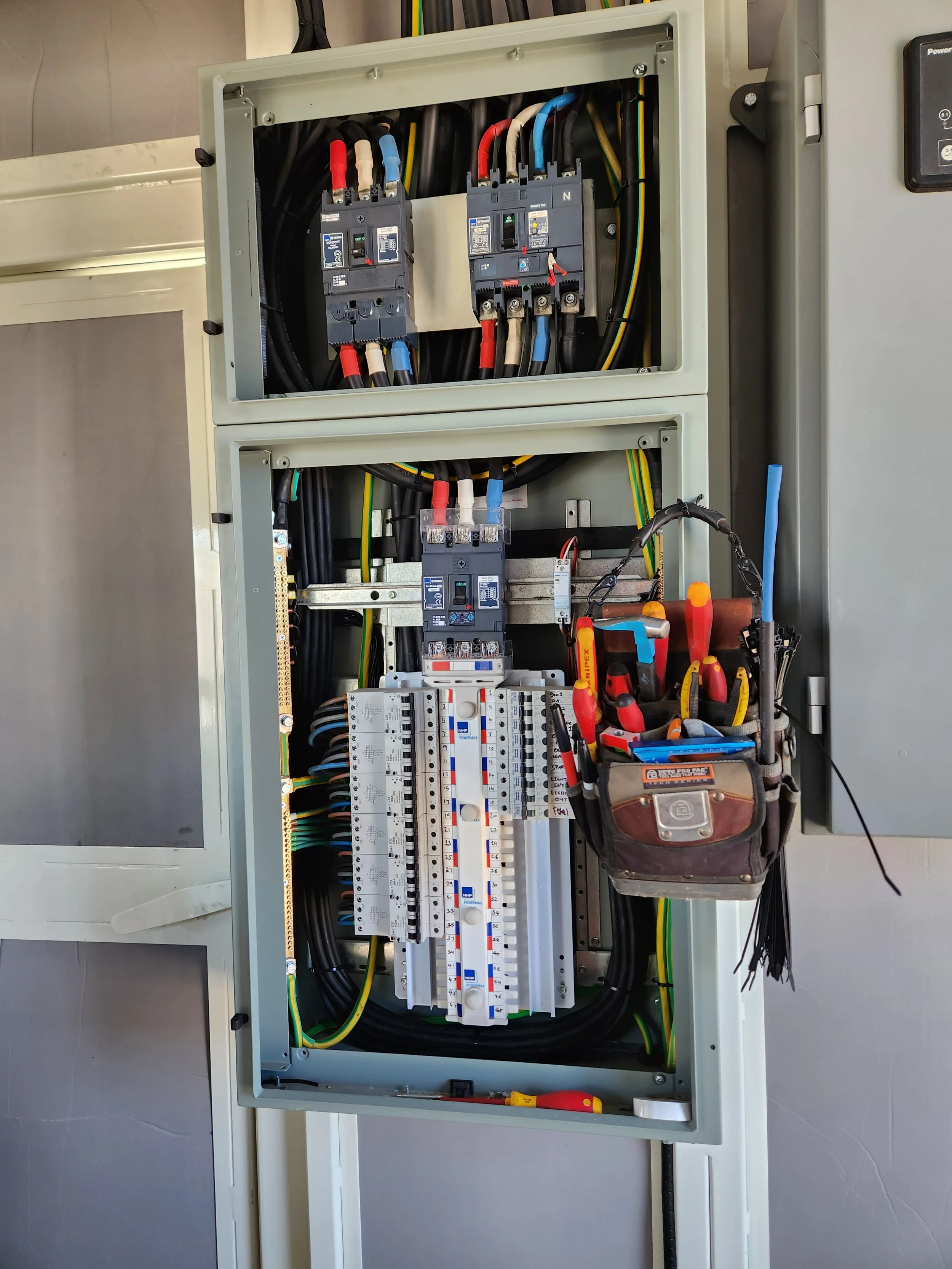 An electrical panel with several circuit breakers, wires, and tools inside a metal enclosure, mounted on a wall.