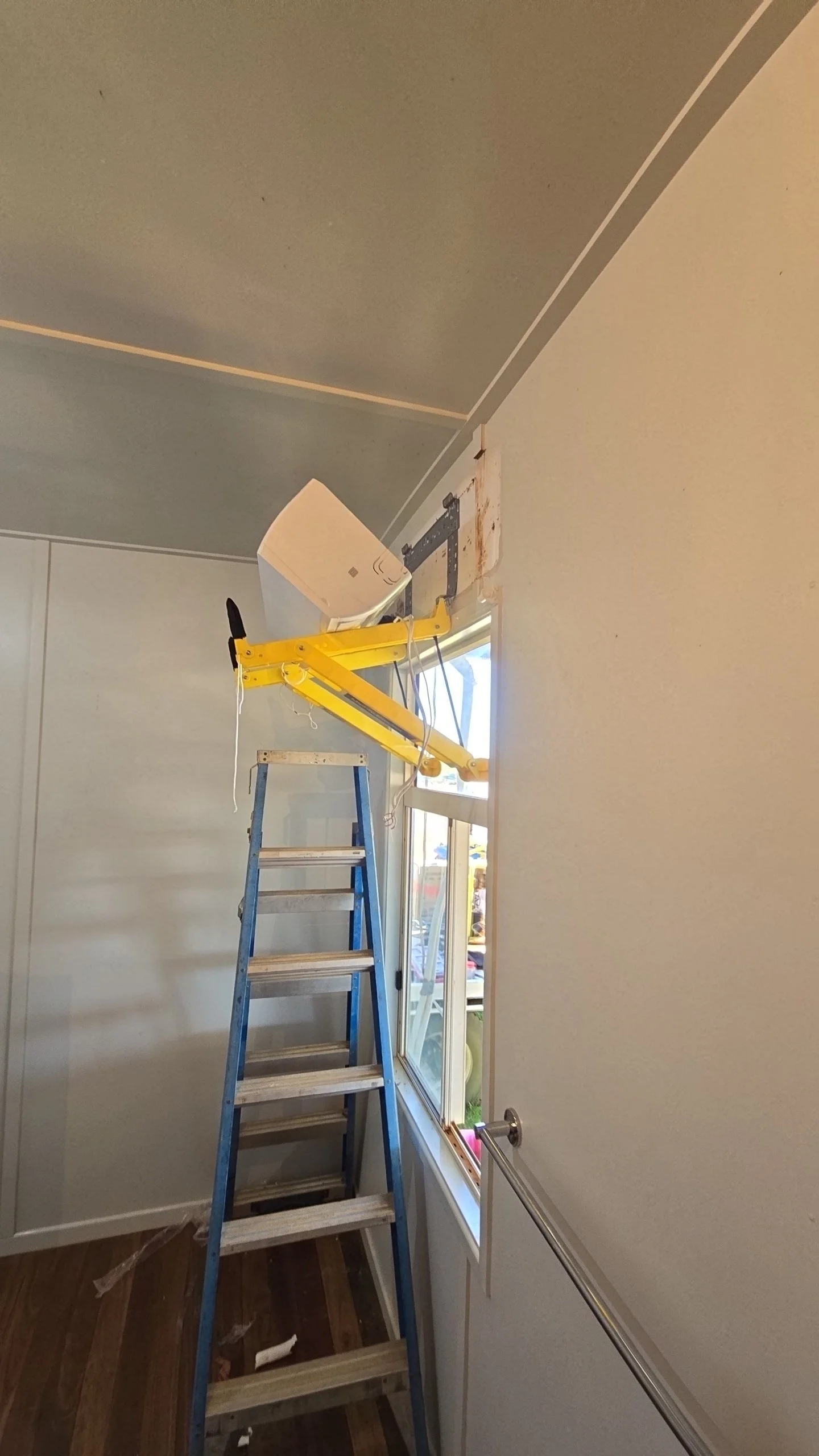 Room under renovation with a blue ladder, yellow tool lift, and a detached window frame, with construction tools and materials on the floor.