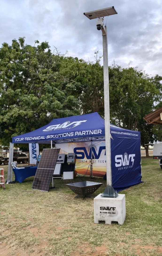 A solar-powered street light with a solar panel at the top, installed on a concrete base labeled 'SWFT'. Behind it, there is a blue tent with the logo and slogan 'Your Technical Solutions Partner' from South West Tech. The tent displays solar energy equipment, and a large solar panel is leaning against the tent.