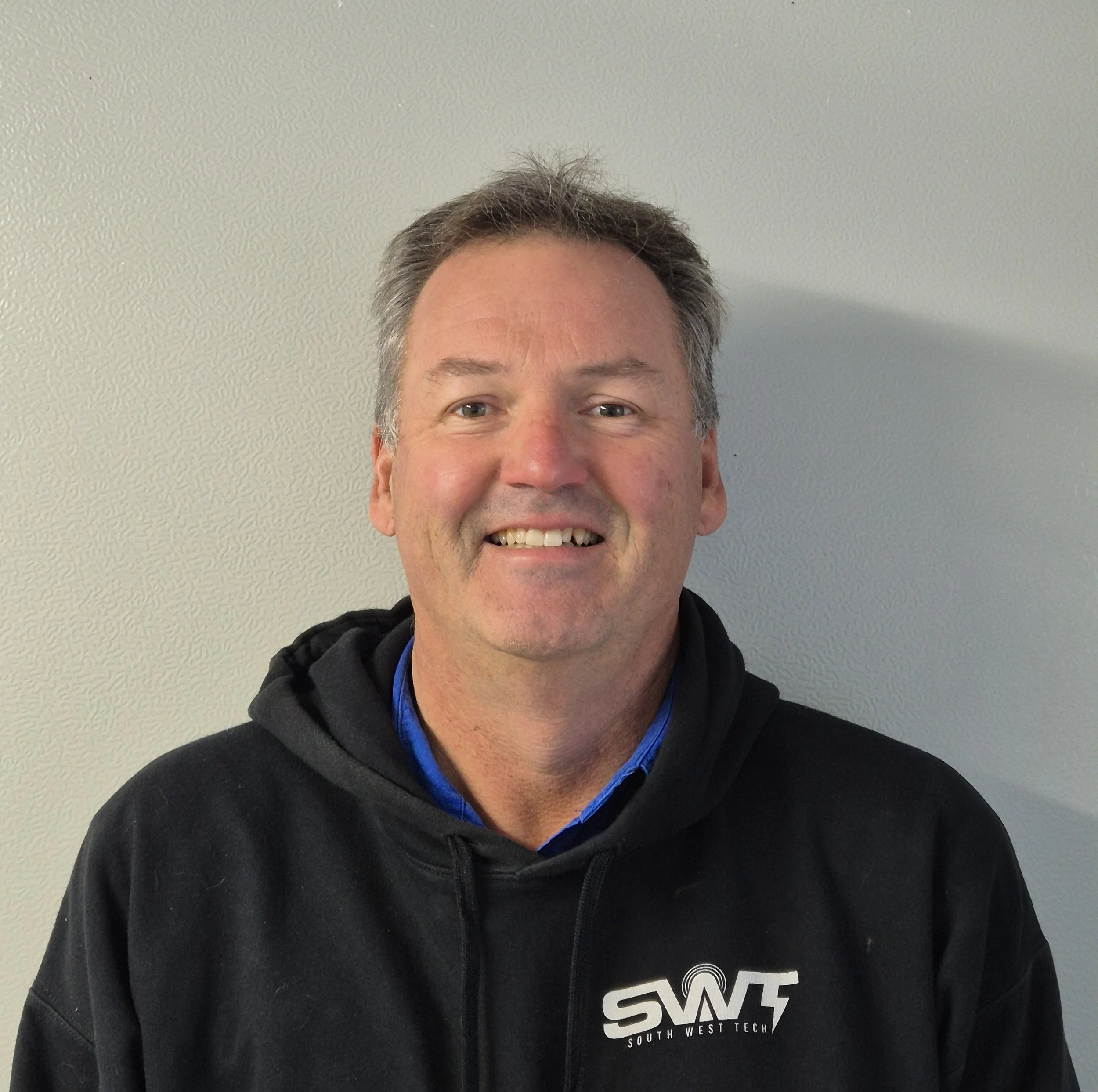 Partial view of a smiling man with gray hair wearing a black hoodie with the words "South West Tech" printed on it, standing against a plain white textured wall.