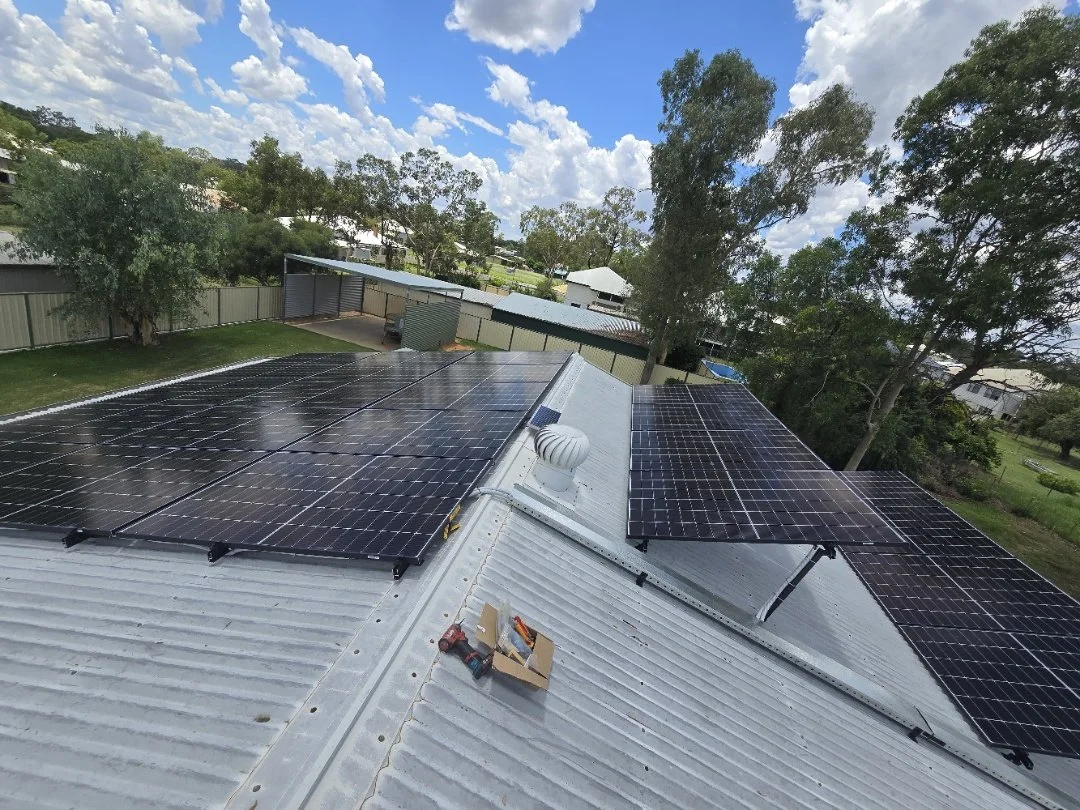 Multiple black solar panels installed on the metal roof of a building, with trees, a fence, and a partly cloudy sky in the background.
